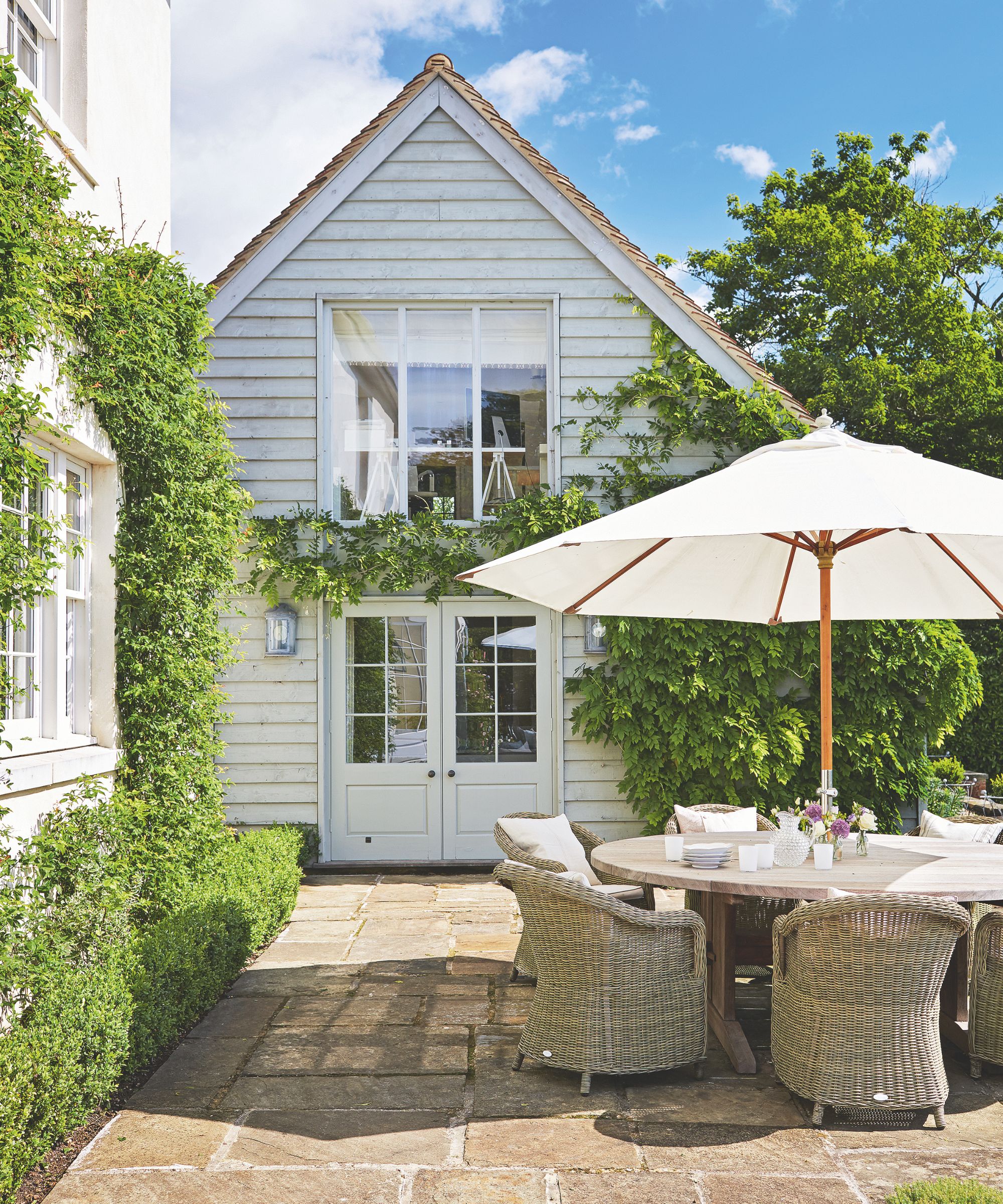 Extérieur d'une maison en bois avec un patio doté d'un mobilier d'extérieur en osier, d'une table à manger en bois, d'un parasol crème et d'un sol carrelé, avec des haies et des buis encadrant la zone.