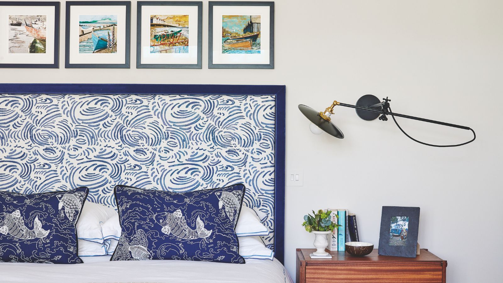 Une chambre blanche avec un lit double à gauche, une tête de lit à motifs bleus et blancs et des coussins bleus à motifs de poissons koi. À droite de la tête de lit se trouve une table de chevet en bois marron avec une petite plante, des livres, un petit bol et une photographie encadrée. Au-dessus se trouve une lampe d’angle murale et au-dessus du lit se trouvent quatre petites œuvres d’art encadrées de manière carrée.