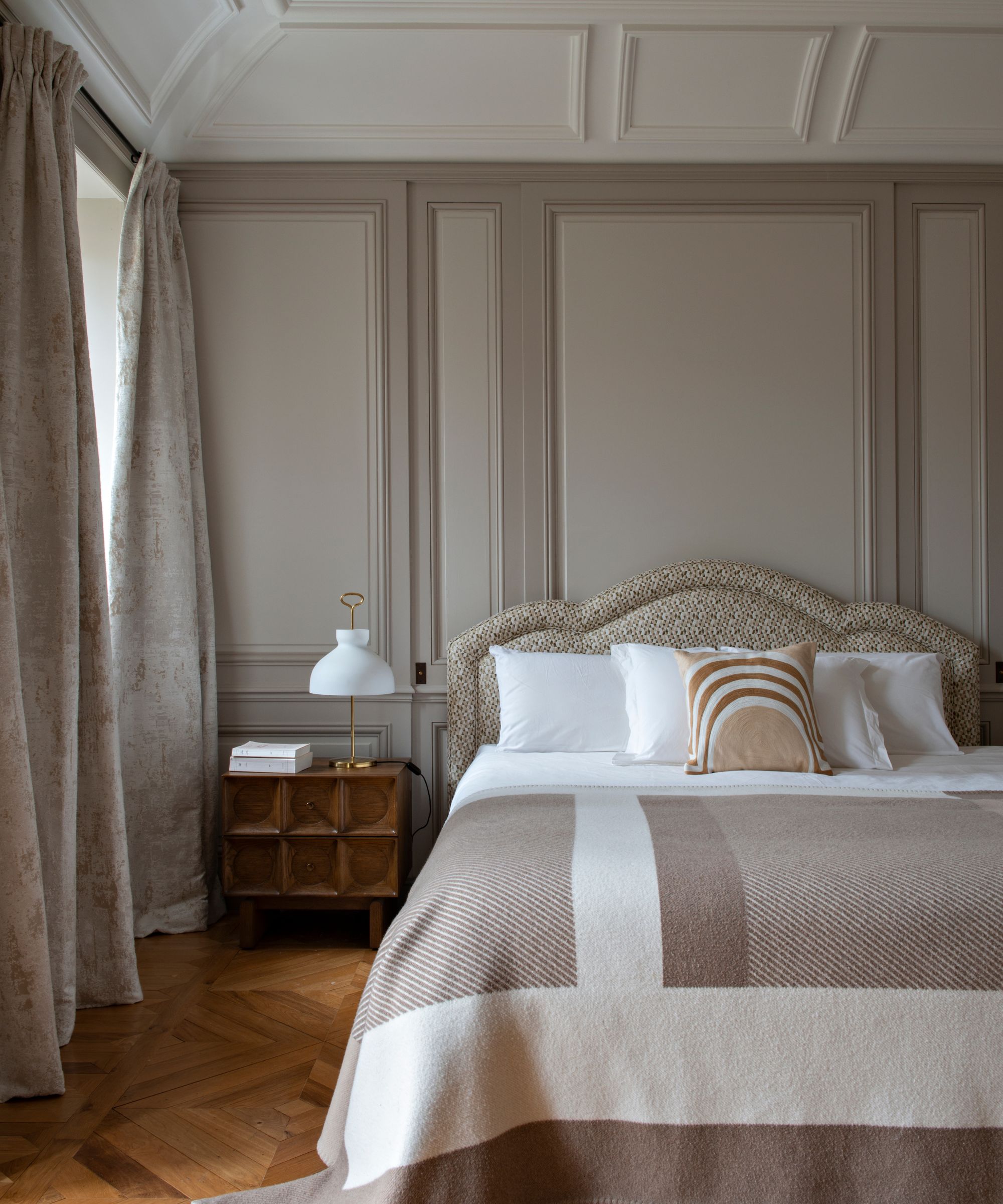 Chambre avec murs peints en Farrow &amp; Ball's Elephant's Breath, parquet au sol, tête de lit rembourrée, draps blancs et couette beige, table de nuit en bois avec lampe en verre sur le dessus.