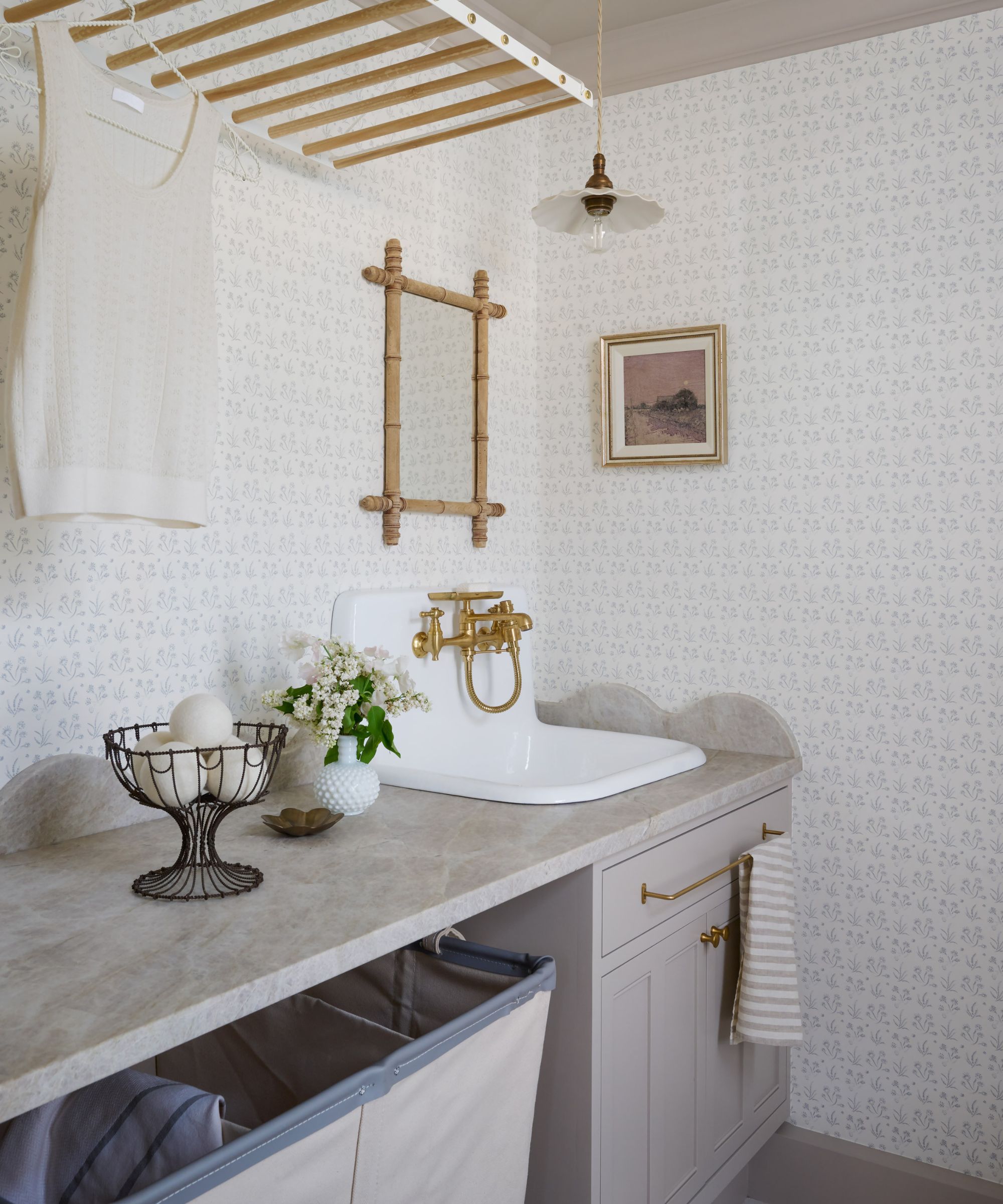 Buanderie avec murs tapissés, Elephant's Breath de Farrow &amp; Armoires peintes en forme de boule, évier en céramique avec robinet doré, panier à linge en dessous et support suspendu en bois au-dessus.
