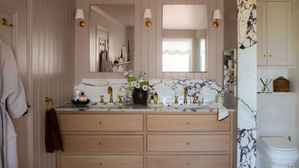 Bathroom with Elephant's Breath by Farrow &amp; Ball painted walls, a double sink vanity unit with marble countertops, wooden drawers, two mirrors and sconces either side.