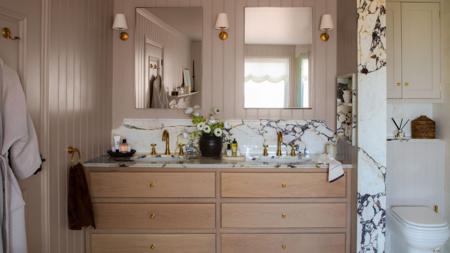 Pourquoi les designers affirment que Elephant’s Breath de Farrow & Ball est la peinture grise la plus chic à utiliser en 2026 – « C’est un neutre pour les personnes qui ne choisissent généralement pas les neutres » Bathroom with Elephant's Breath by Farrow & Ball painted walls, a double sink vanity unit with marble countertops, wooden drawers, two mirrors and sconces either side.