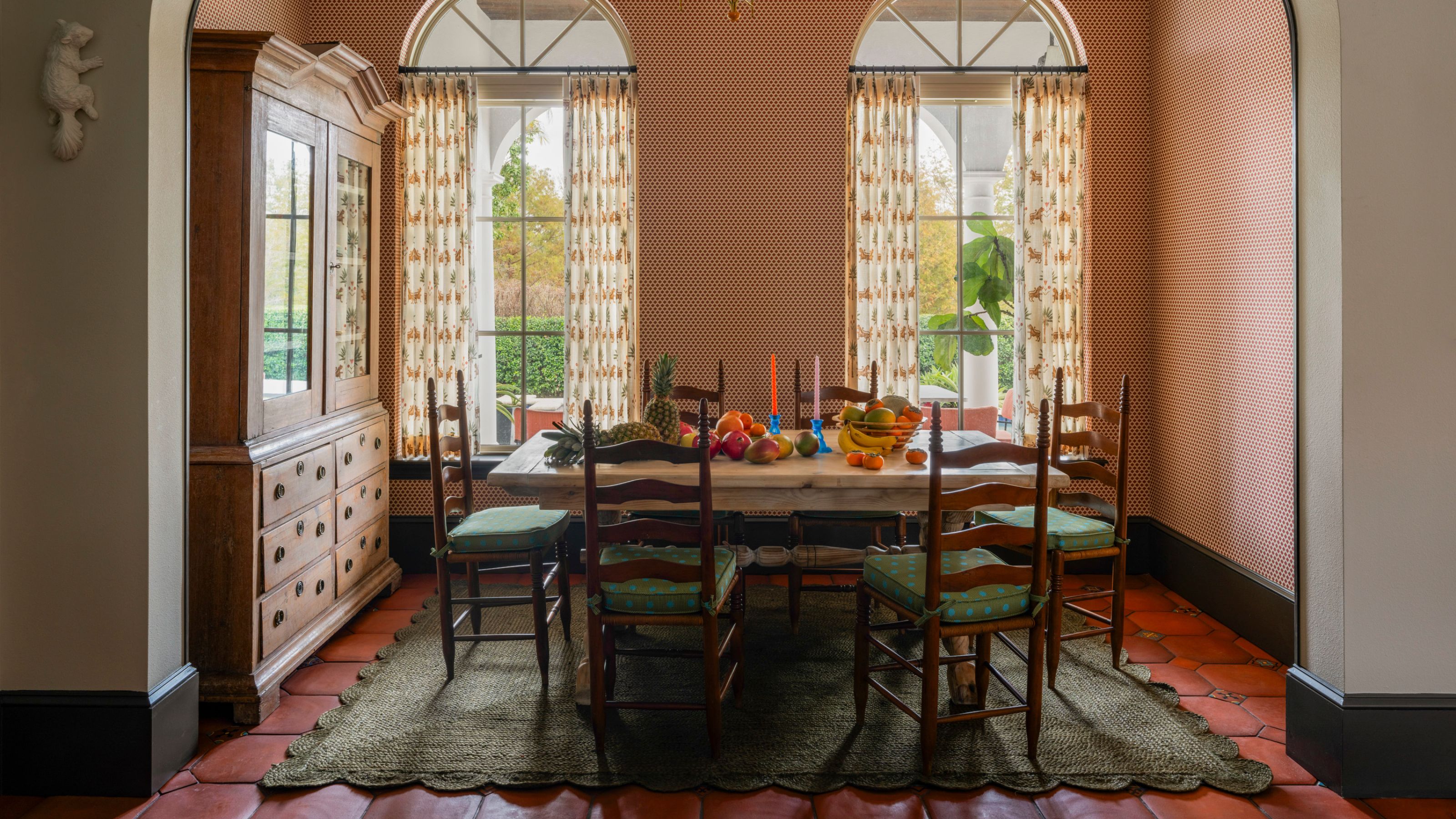 une salle à manger ensoleillée dans une maison de Miami avec de grandes portes-fenêtres cintrées, des murs tapissés, des sols en terre cuite, un tapis vert festonné et des meubles anciens