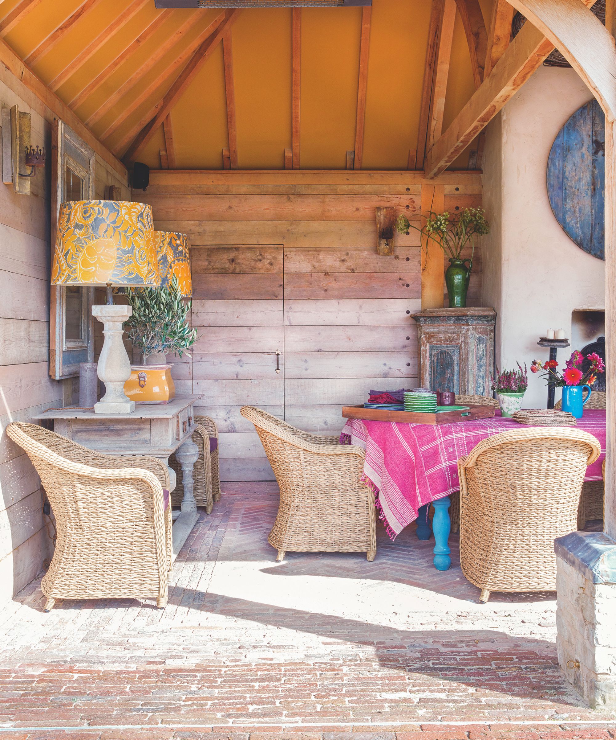 Grange extérieure aménagée avec sol en brique, fauteuils en rotin disséminés autour d'une table à manger bleue avec une nappe rose et deux grandes lampes de table jaunes sur une console.