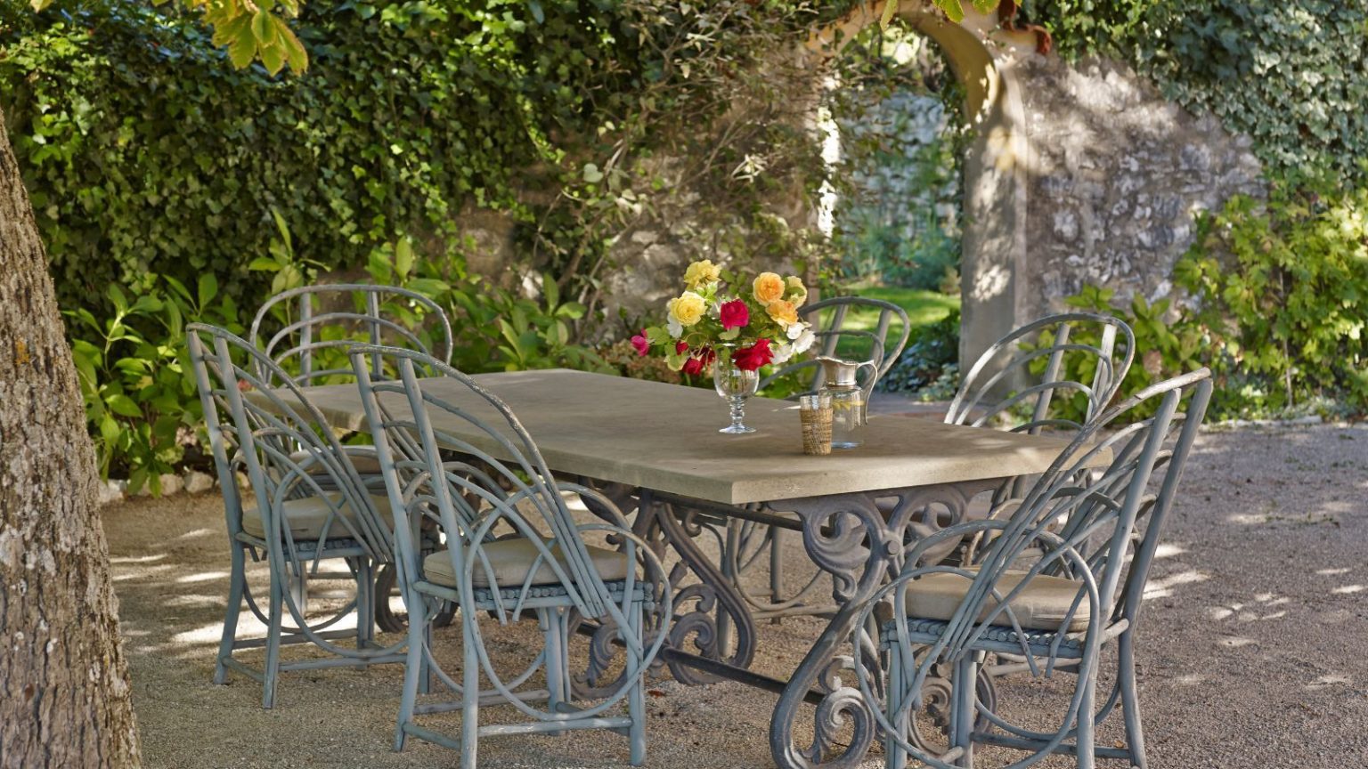 French garden with a brick arched way covered in ivy, and an antique wrought iron long dining table with vintage cane chairs underneath.