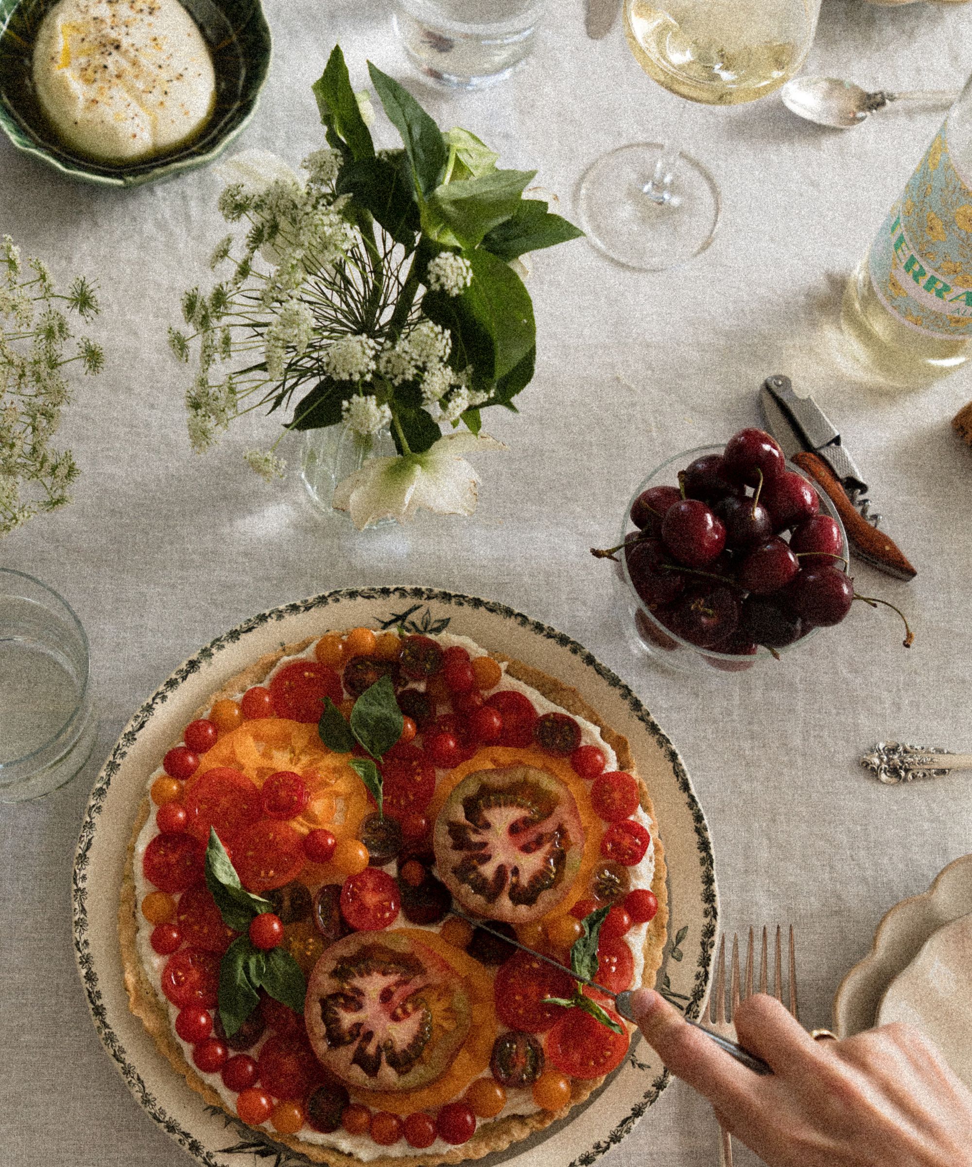 Décoration de table extérieure avec une nappe en lin, un vase à boutons rempli de fleurs blanches fraîches, une assiette en porcelaine verte et blanche avec une tarte aux tomates, un bol de cerises et des verres à vin