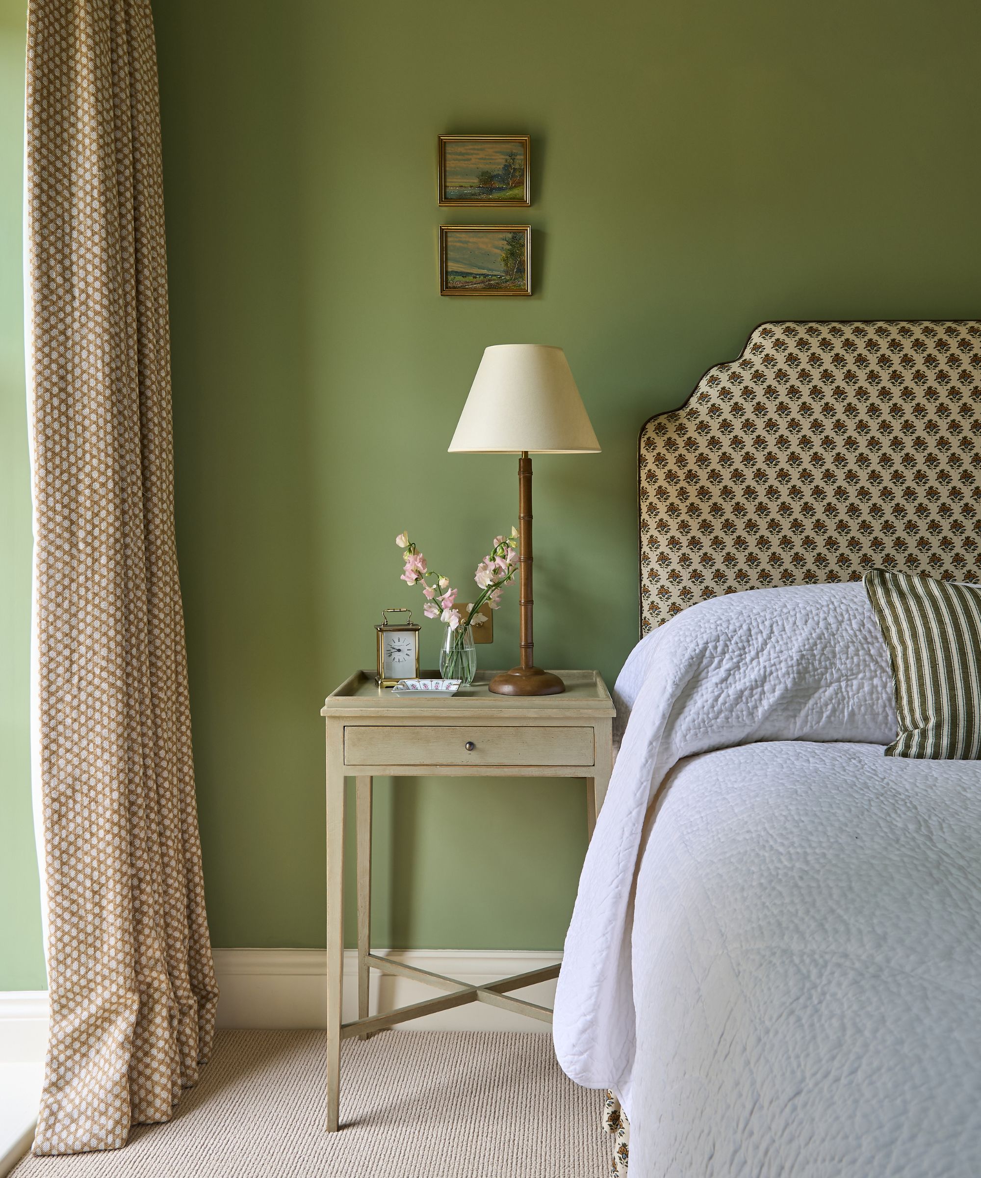 Chambre traditionnelle avec des murs vert olive doux, une tête de lit rembourrée à motifs, une table de chevet en bois clair ornée de fleurs fraîches et une lampe de table classique.