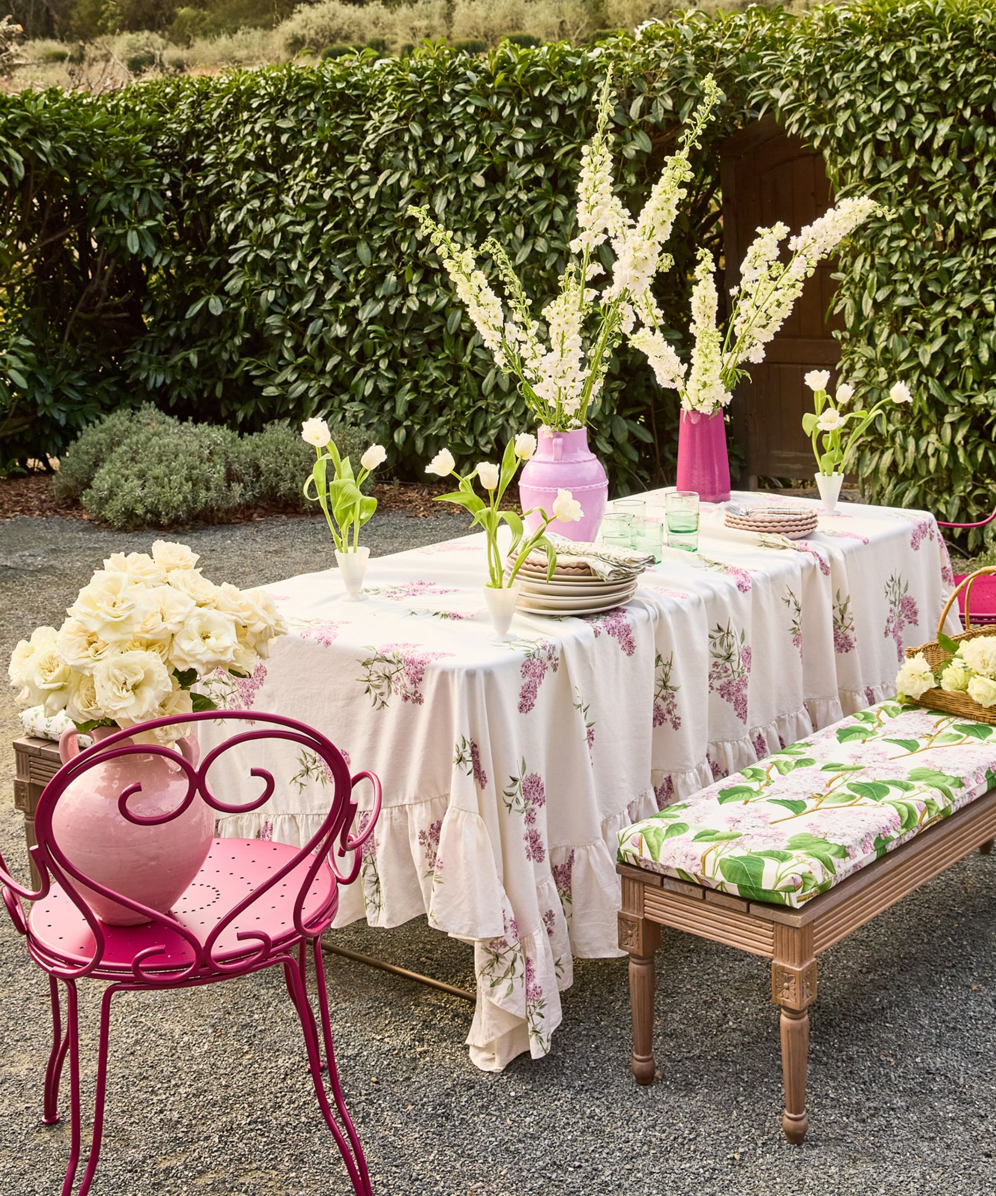 une table extérieure avec une nappe fleurie, une chaise en métal rouge, un banc avec un coussin fleuri et des fleurs décoratives le long de la table