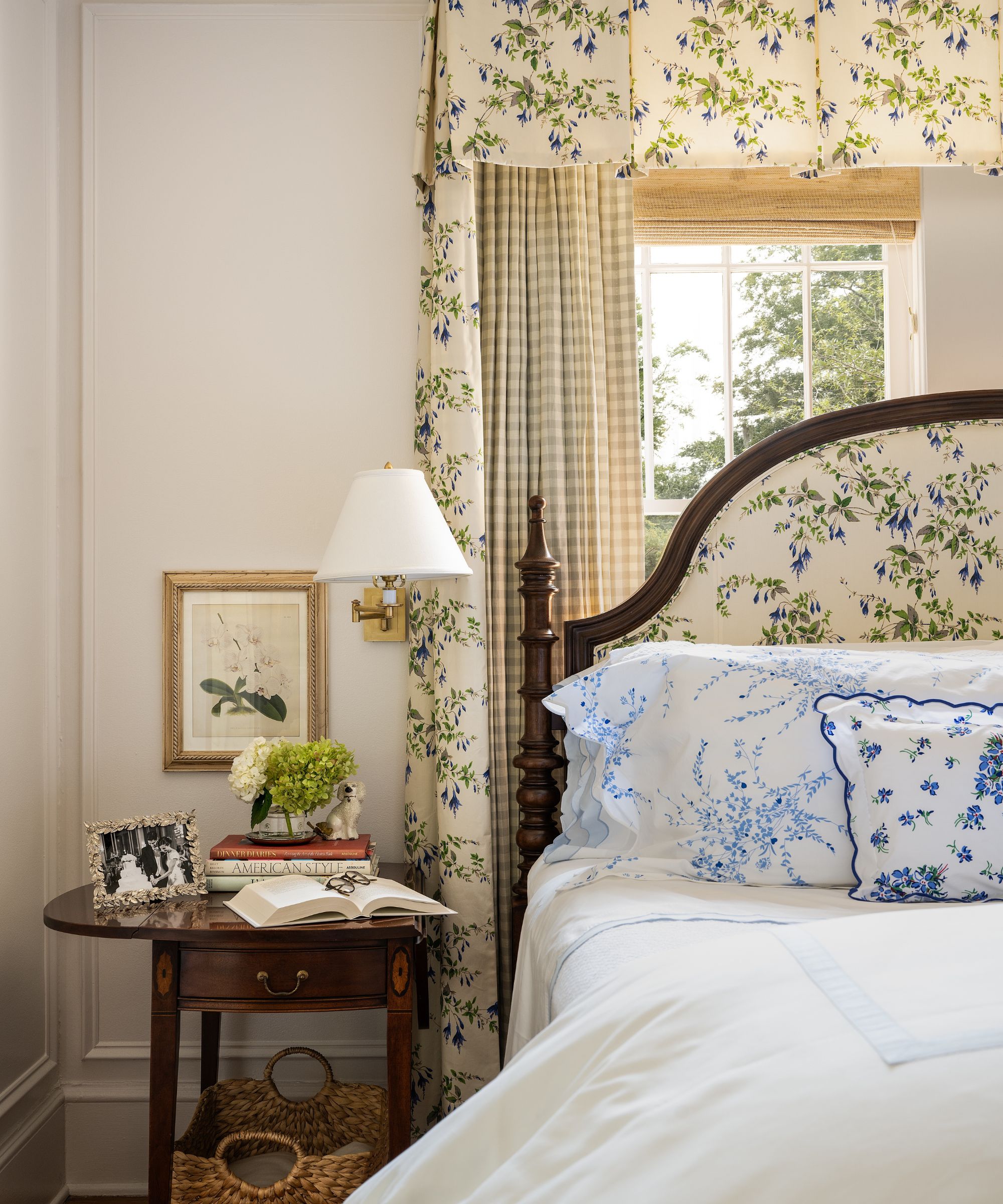 Chambre avec des murs crème, une fenêtre avec un store romain en bambou, un ciel de lit fleuri avec des rideaux en vichy, un lit en acajou recouvert d'un imprimé floral et une table de nuit avec des livres et une applique au-dessus.