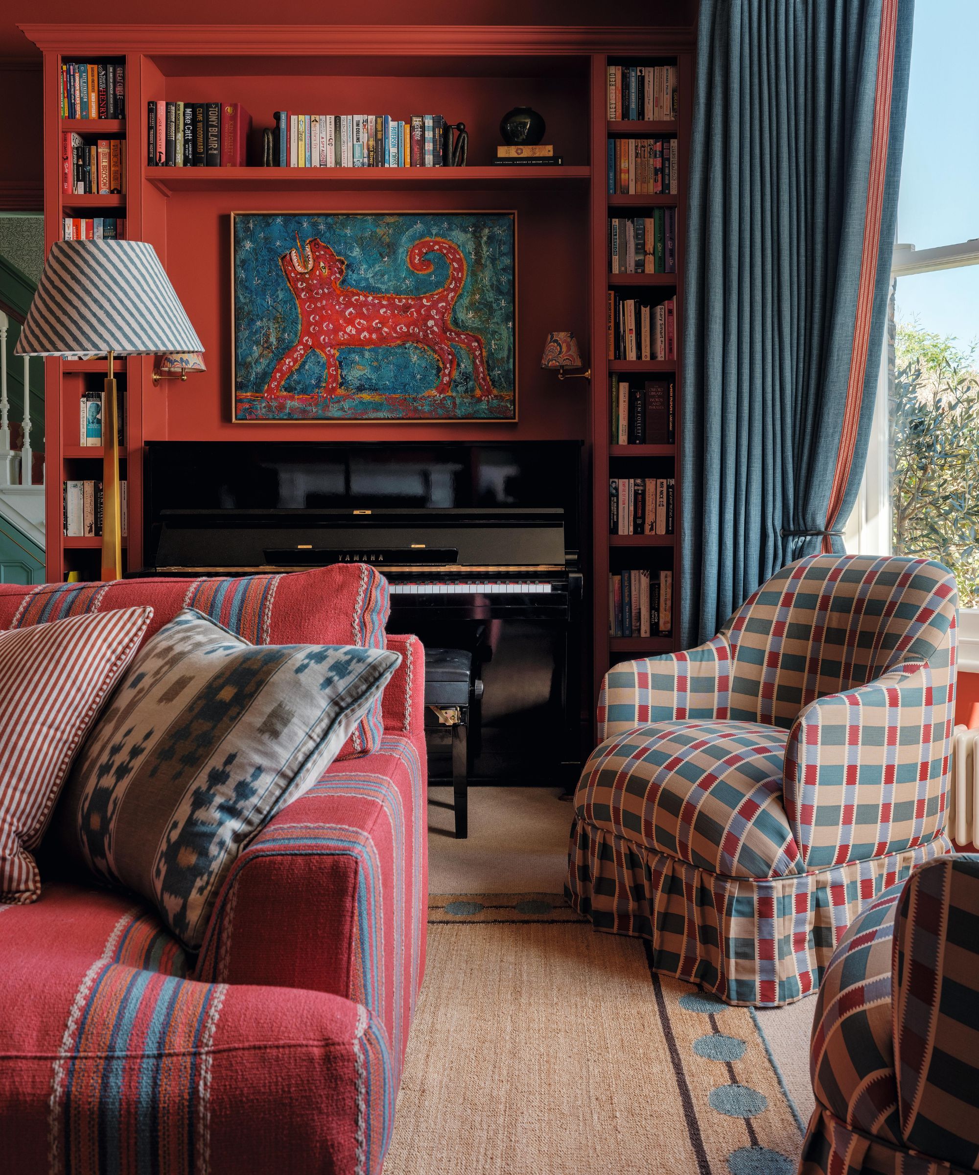 Un salon peint en rouge avec une grande fenêtre habillée de rideaux bleus, un espace bibliothèque sur mesure construit autour d'un piano, un canapé rouge à rayures et une paire de fauteuils à carreaux