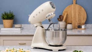 A cream retro style SMEG stand mixer in a blue kitchen, on white marble work tops.