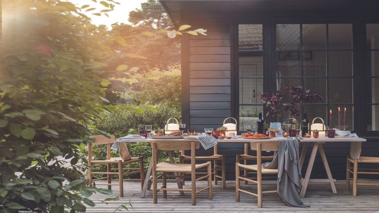 La vente de liquidation de jardin de QVC est arrivée – Préparez votre jardin pour l’été avec les meilleures offres à acheter dès maintenant A wood panelled patio in front of a wood panelled home with a large tablescaped dining table, wooden chairs, and a large bush in the left foreground.