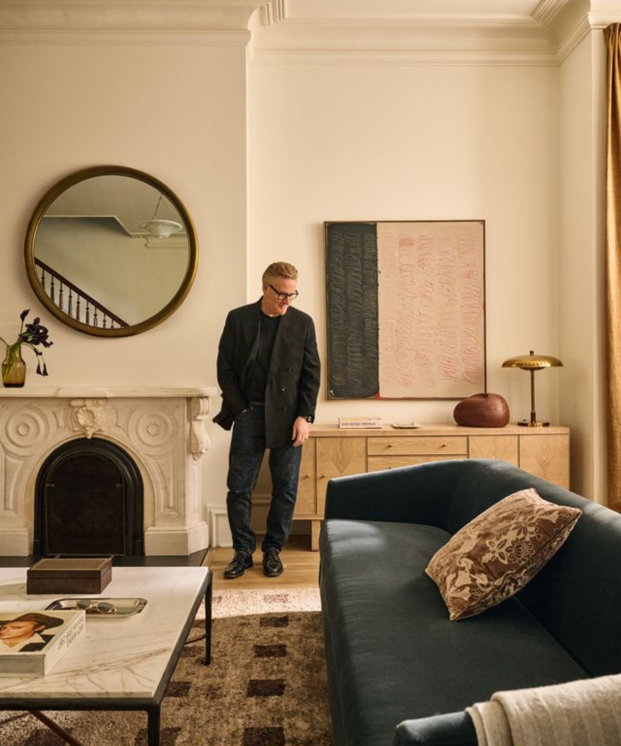 un homme debout dans un salon avec un canapé bleu, une table basse en marbre, une cheminée décorative et un miroir accroché au mur