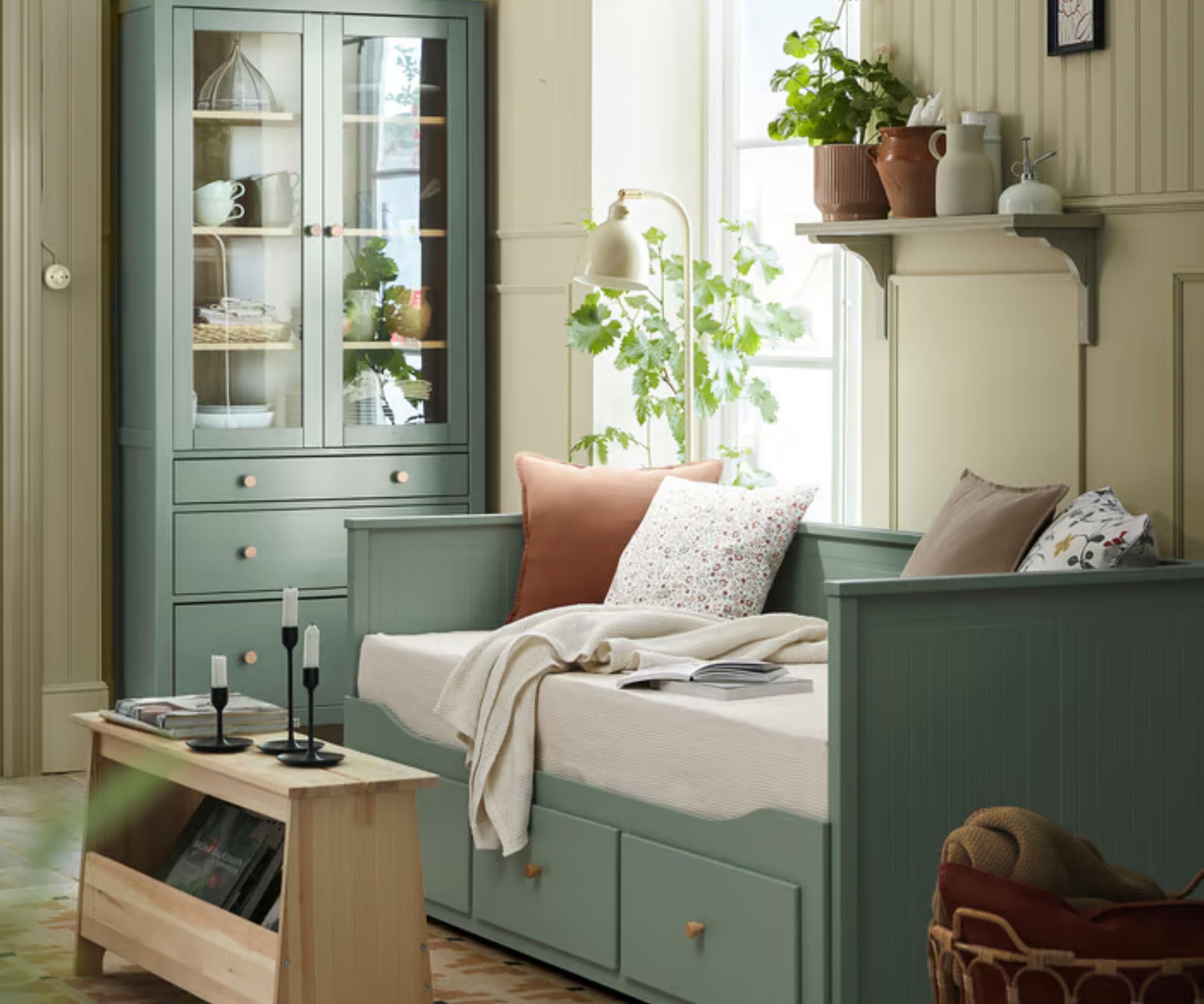 Lit HEMNES IKEA vert sauge avec des coussins neutres, une table d'appoint en bois avec des chandeliers noirs, une commode peinte en vert sauge avec des céramiques et de la vaisselle et un mur lambrissé peint en crème.