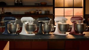 Five kitchenaid artisan plus stand mixers with stainless steel bowls on the dark counter of a red kitchen, in half light.