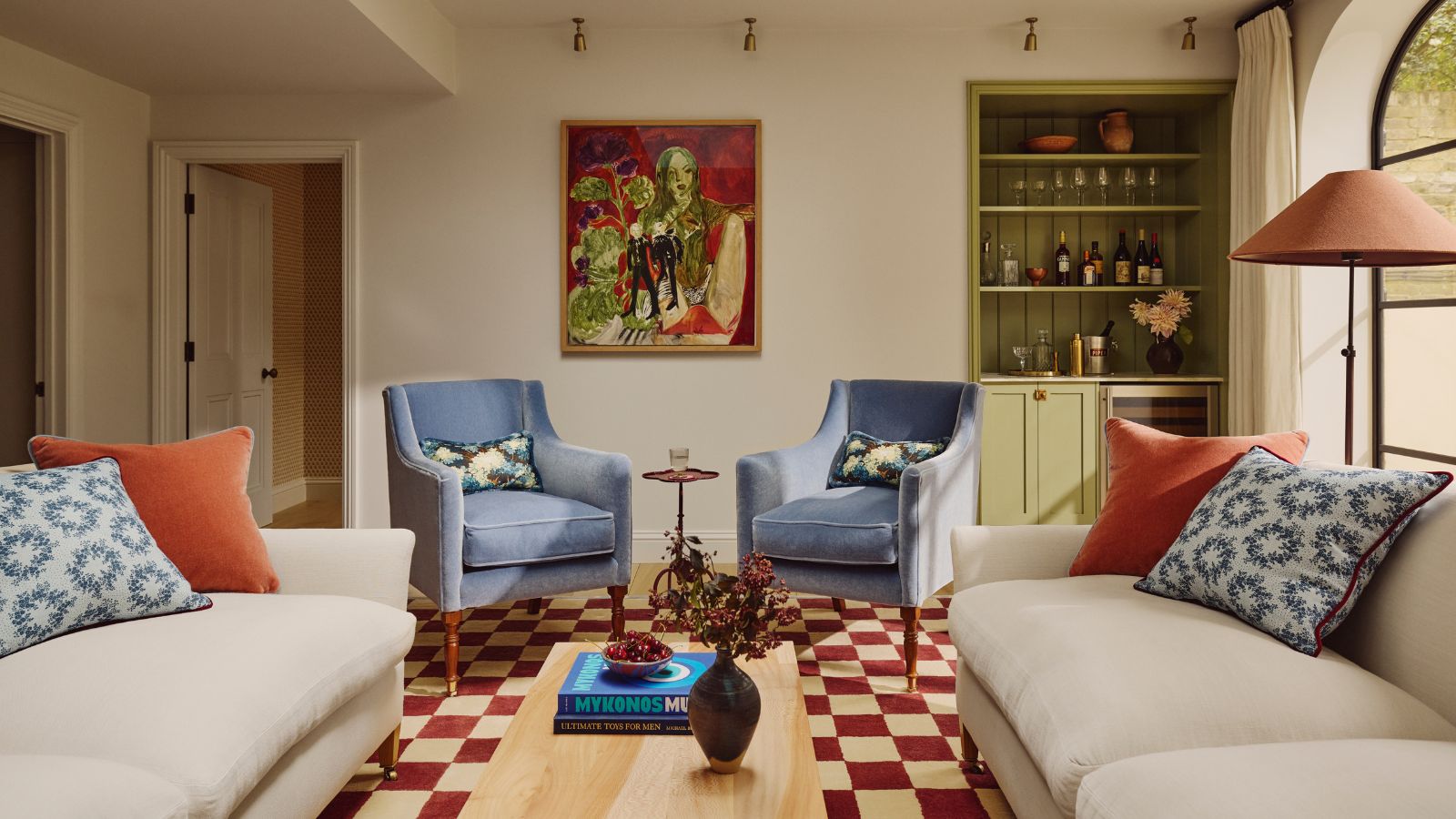 un salon avec un tapis à carreaux, deux fauteuils d'appoint bleus, deux canapés blancs avec des oreillers sur le dessus, un bar vert intégré sur le côté droit et des œuvres d'art abstraites au mur
