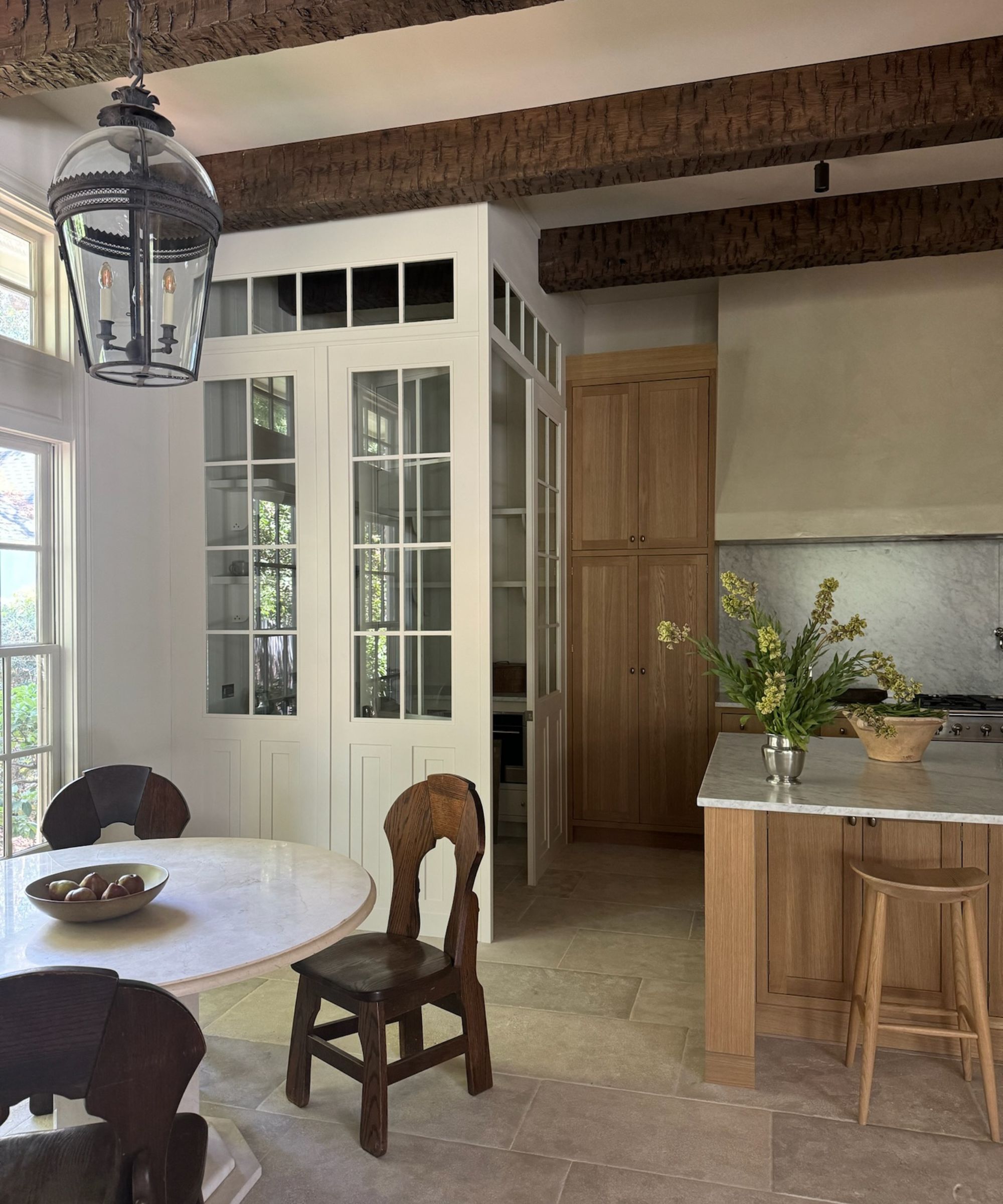 Cuisine neutre avec armoires en bois clair et tabourets de bar, sol en pierre, table à manger ronde en marbre avec chaises en bois sombre. Vase de fleurs sur l'îlot de cuisine.