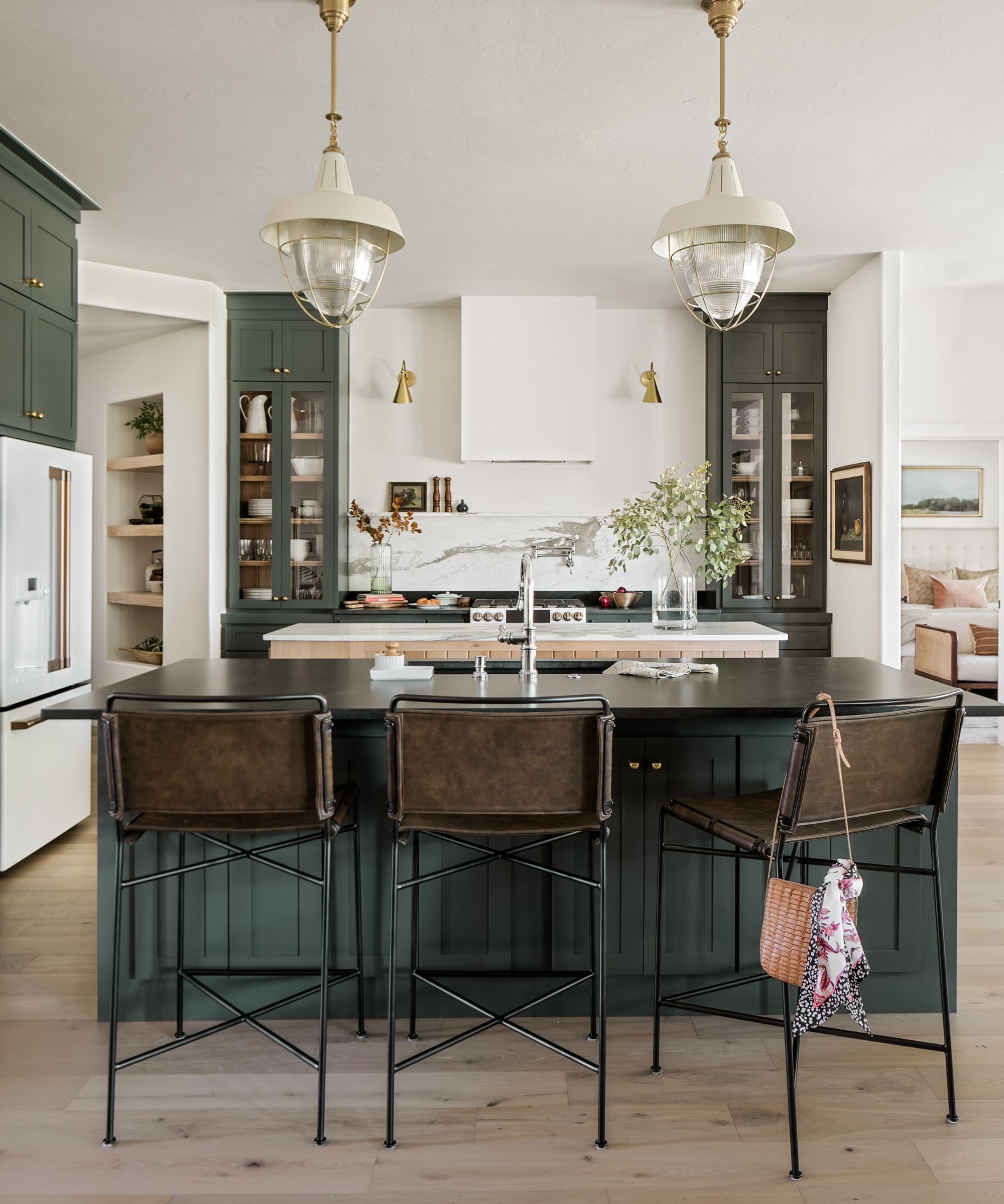 Cuisine avec armoires et îlot peints Pewter Green de Sherwin-Williams, dosseret en marbre, plancher en bois et murs blancs, avec deux pendentifs crème et or suspendus au plafond.