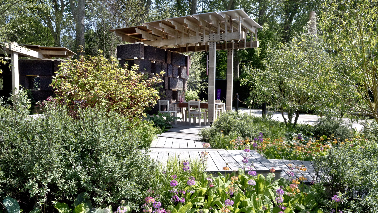 Un jardin d'exposition avec arbustes et primevères bordé d'une passerelle en bois et d'une grande pergola avec coin repas