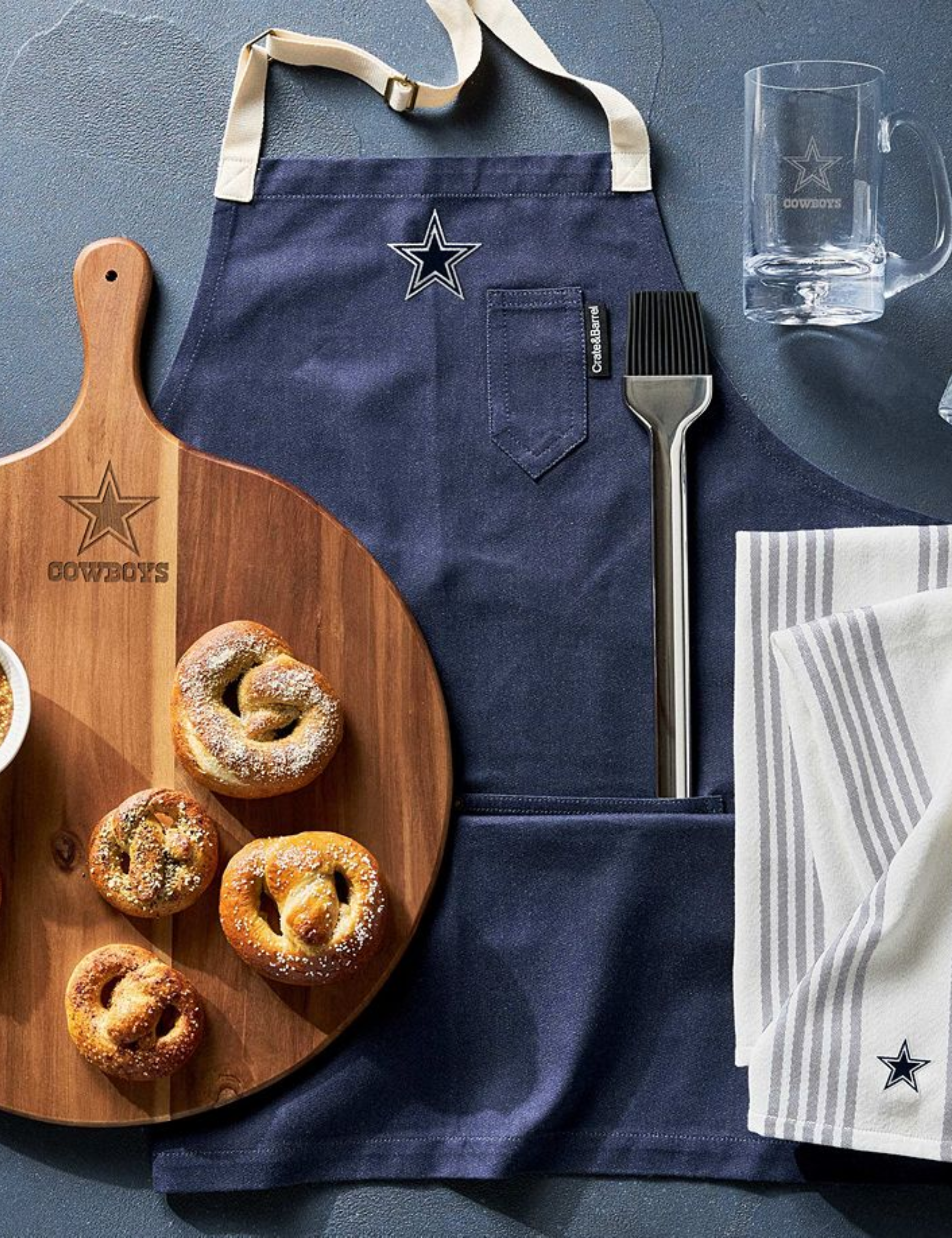 Caisse et amp;amp; Planche de service en bois de marque Barrel x NFL avec bretzels moelleux, tablier, outil de barbecue, chope à bière et torchons photographiés depuis une vue à vol d'oiseau.