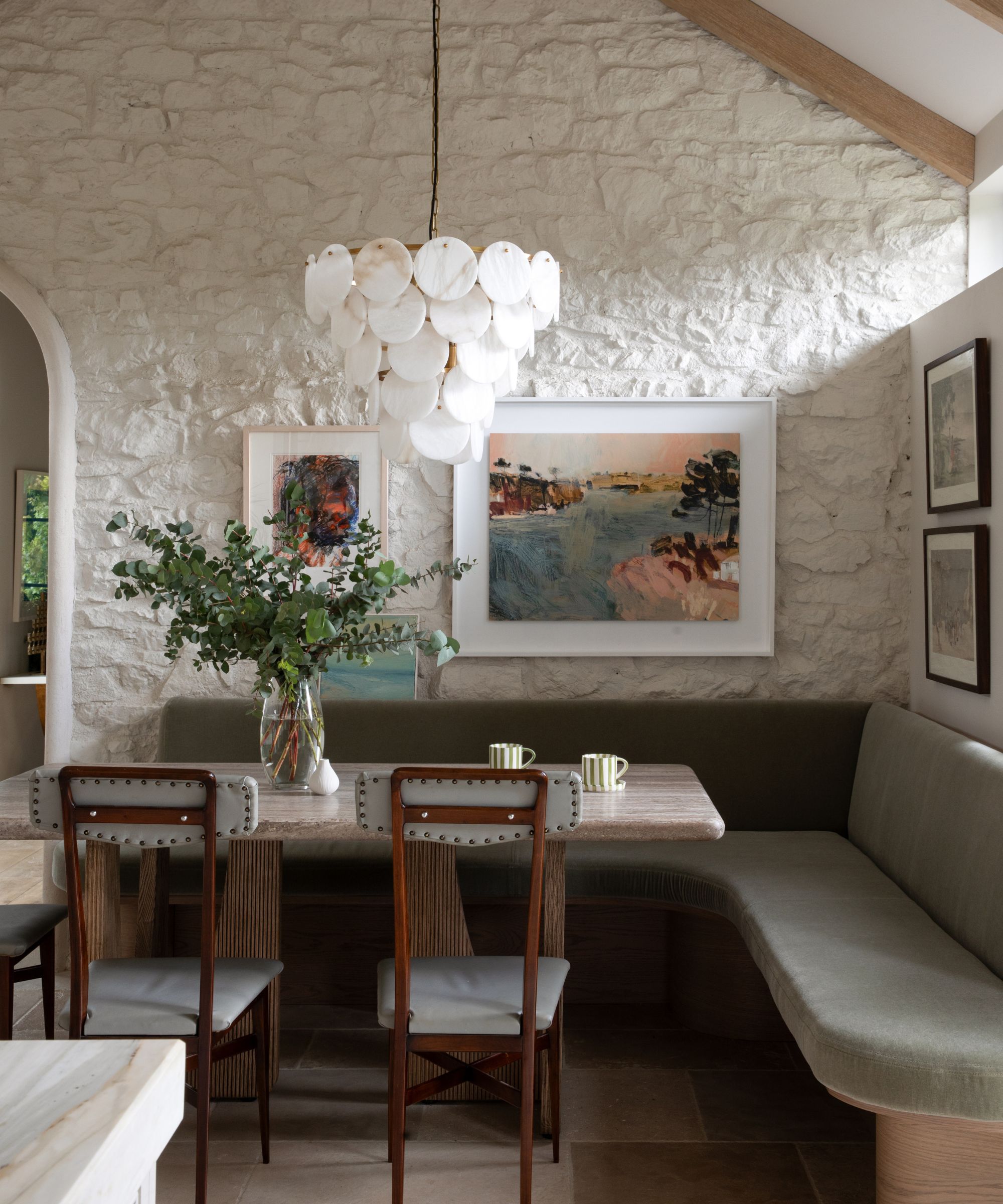 Un coin salon de cuisine avec une banquette verte en forme de L, une table en bois avec un plateau en marbre et des murs en pierres apparentes lavés en blanc et recouverts d'œuvres d'art vintage