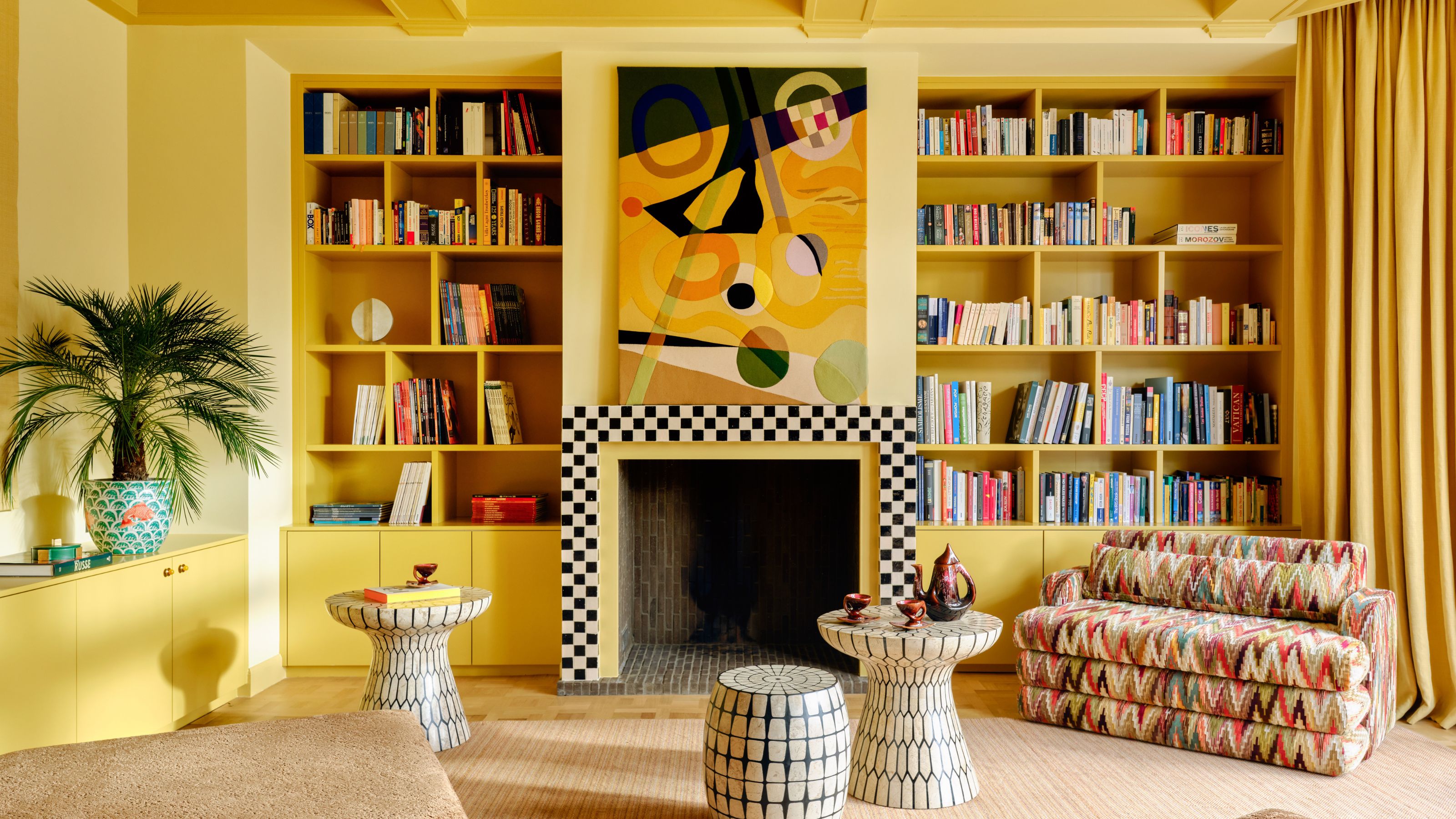 un salon peint en jaune vif avec un foyer carrelé en damier, des bibliothèques et des meubles et œuvres d'art éclectiques