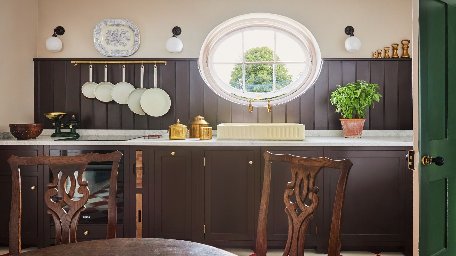 Cuisine rustique avec boiseries marron foncé, comptoirs en marbre blanc, évier de couleur crème sous une grande fenêtre ovale et chaises de salle à manger en bois au premier plan