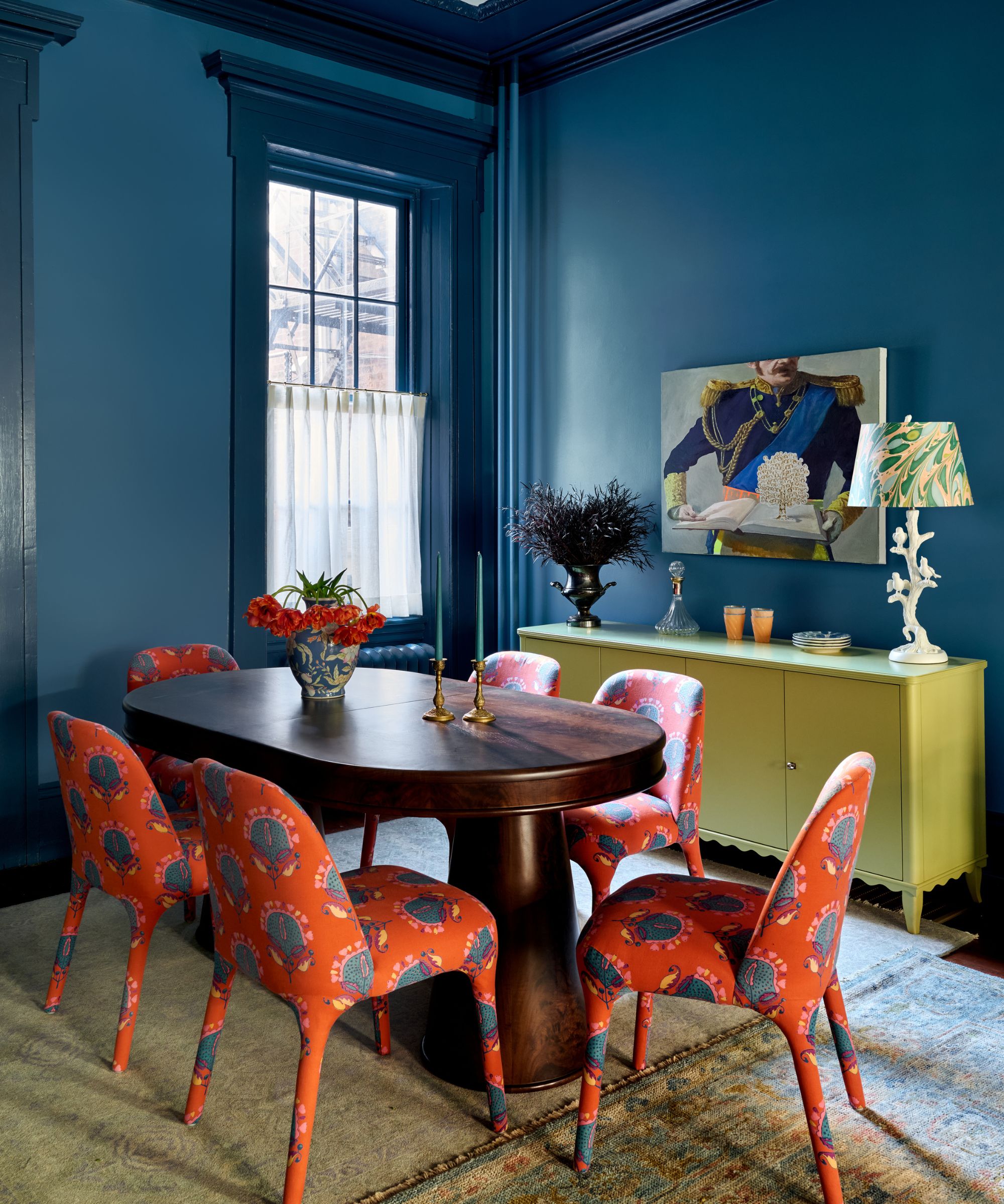 Salle à manger avec des murs bleu sarcelle profond, une table ovale en bois sombre, six chaises motif cachemire corail clair et une crédence vert olive.