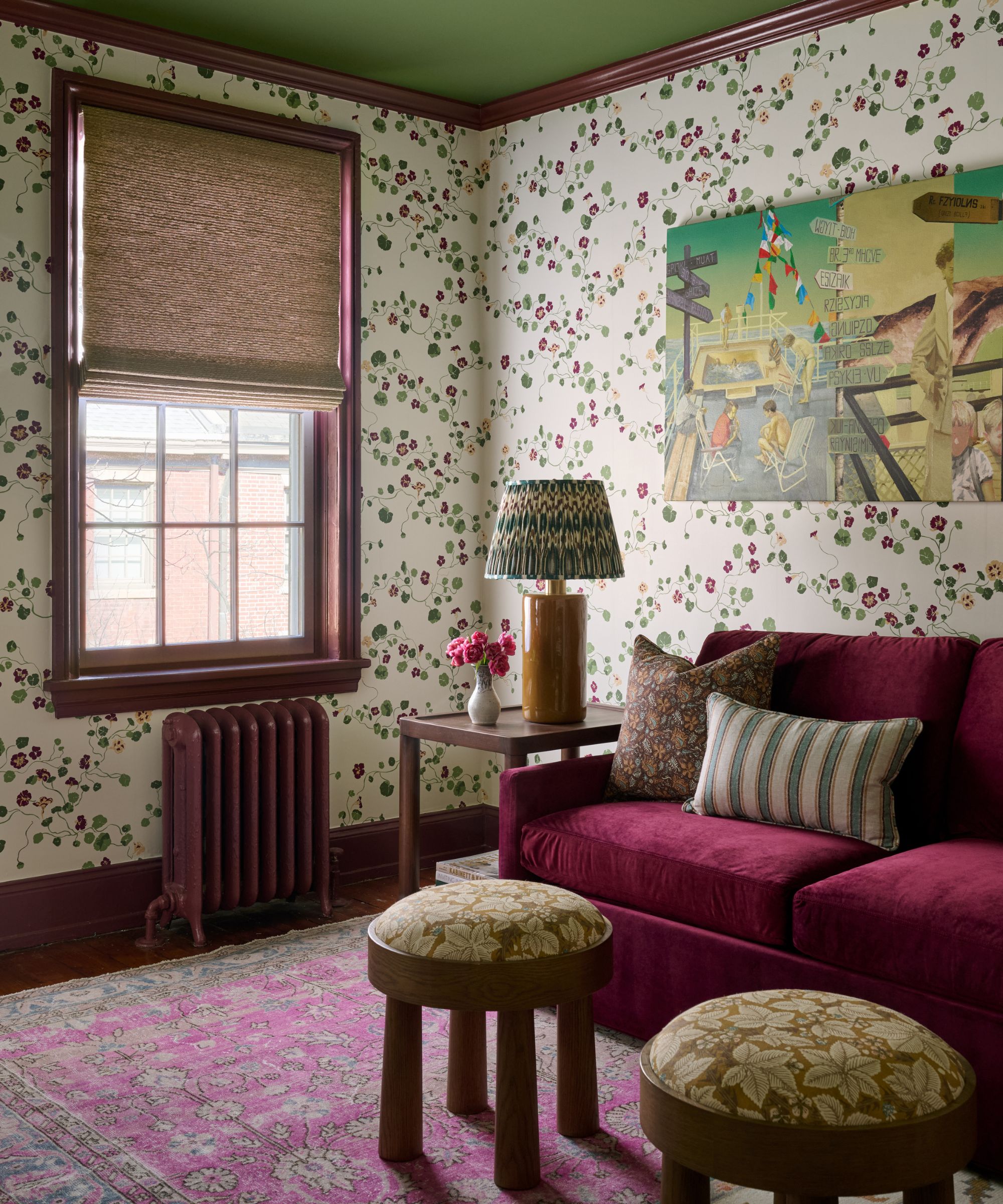 Coin salon avec papier peint fleuri, canapé en velours bordeaux, tapis à motifs roses et deux petits tabourets ronds en bois.