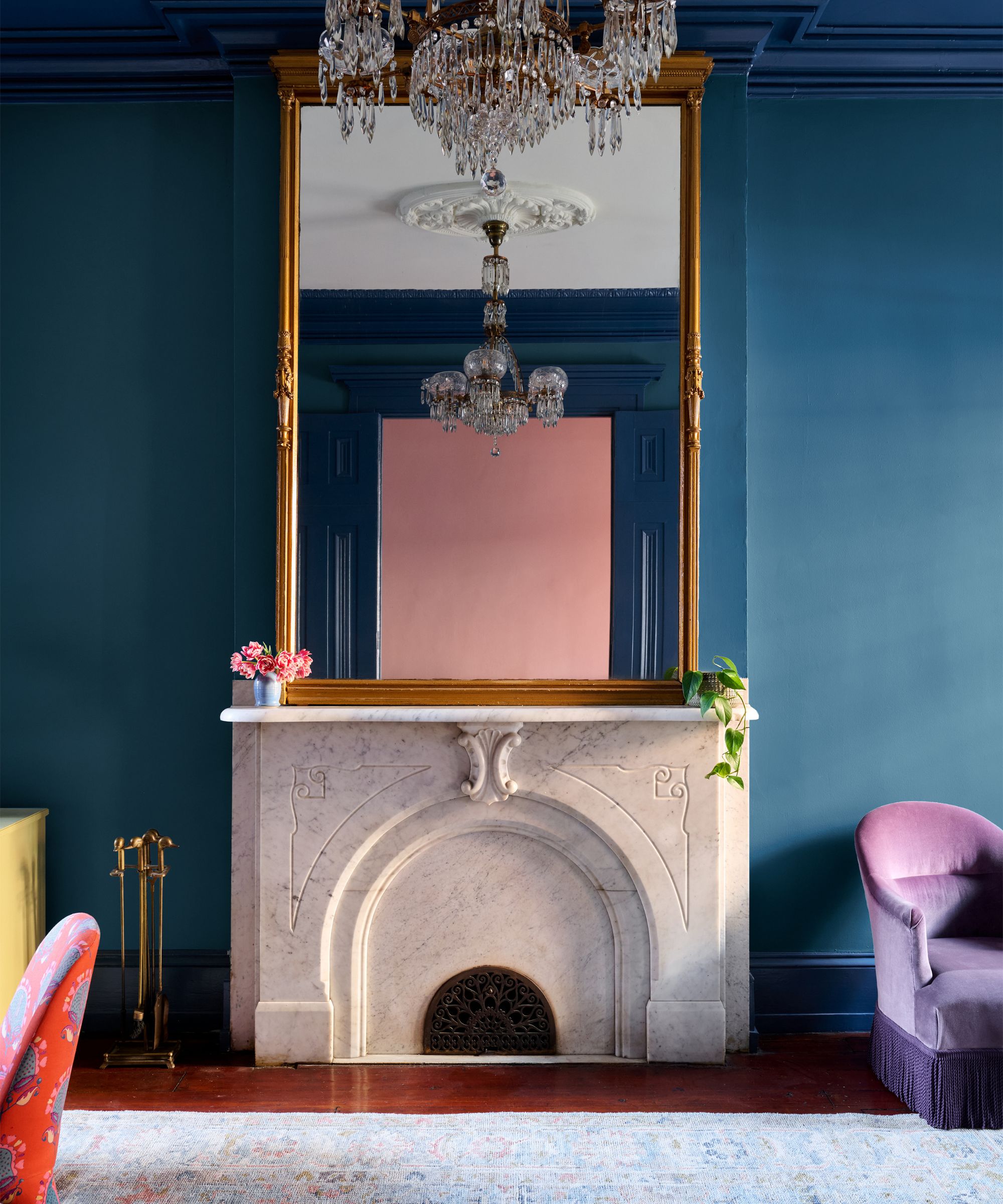 Cheminée en marbre blanc contre un mur bleu sarcelle, avec un grand miroir doré reflétant un mur rose et un lustre en cristal.