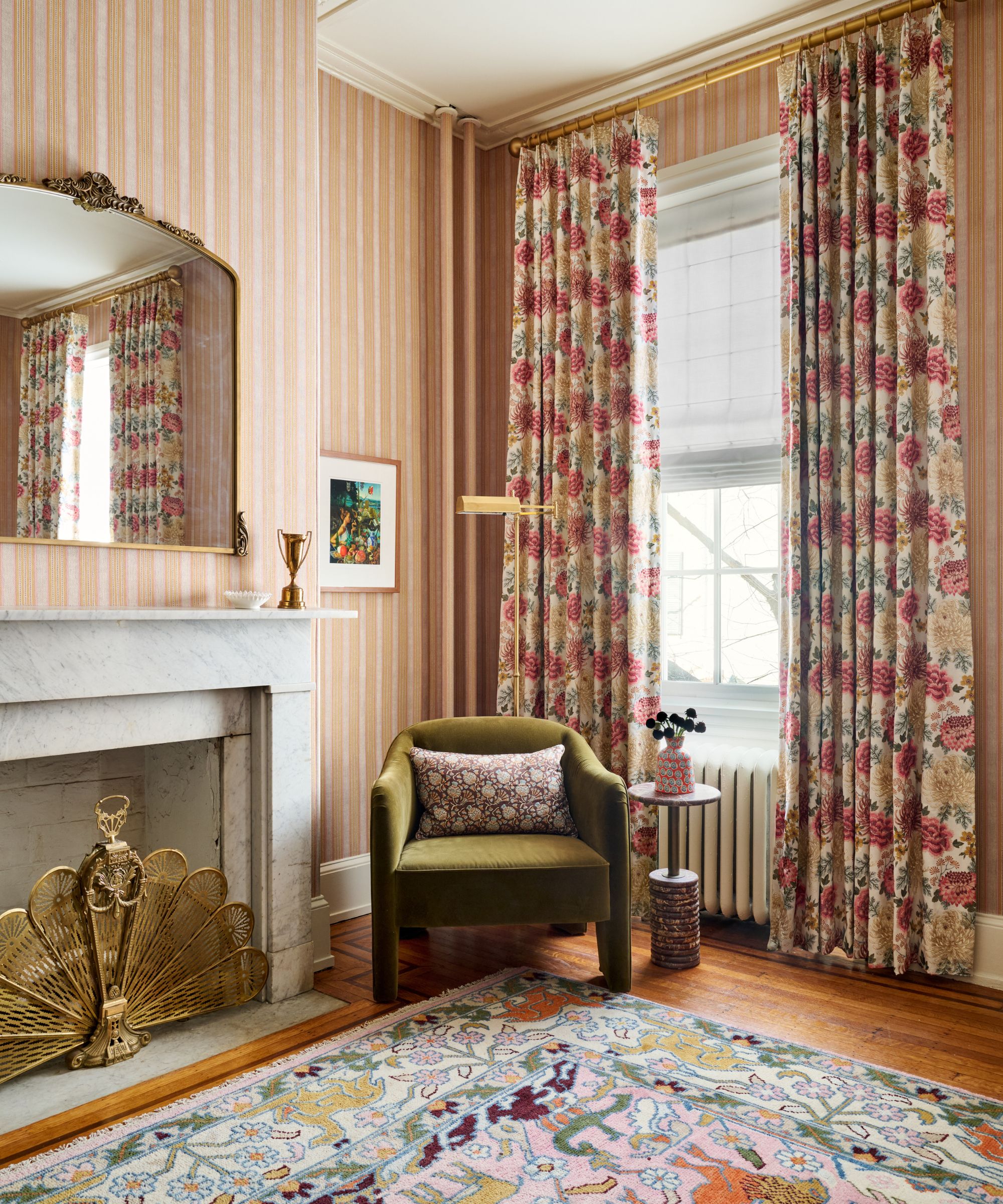 Coin chambre avec cheminée en marbre, miroir en arc doré, fauteuil en velours vert mousse et rideaux fleuris rose vif.