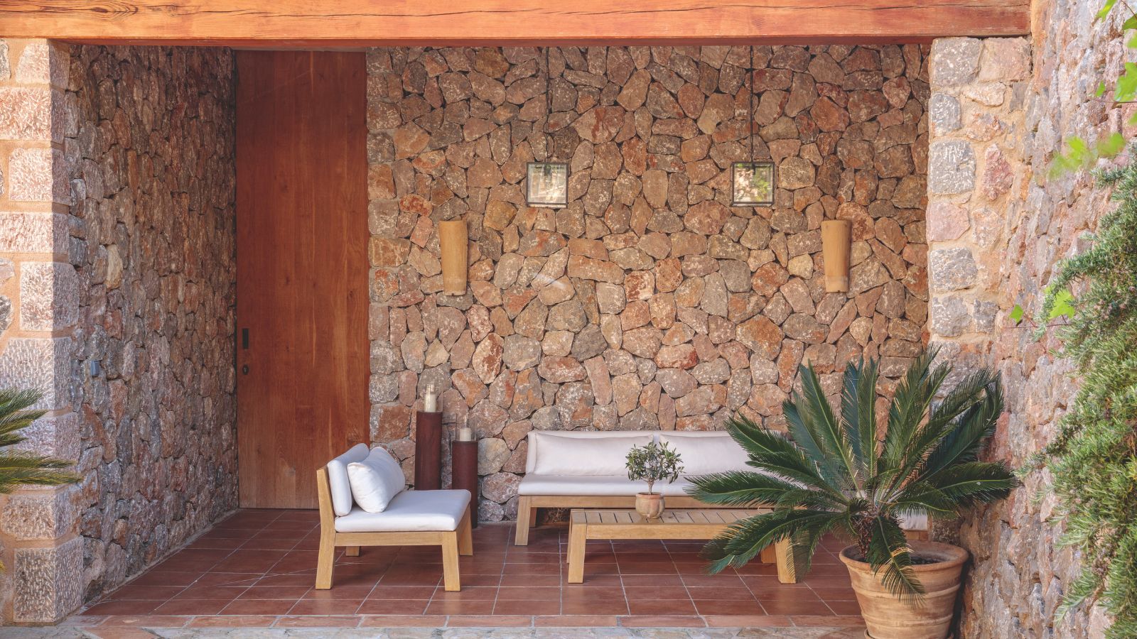 Patio rustique avec un canapé et une chaise d'extérieur blancs et en bois, une table basse en bois, un palmier en pot, des murs en briques, des lanternes en verre suspendues au plafond et un sol carrelé en terre cuite.