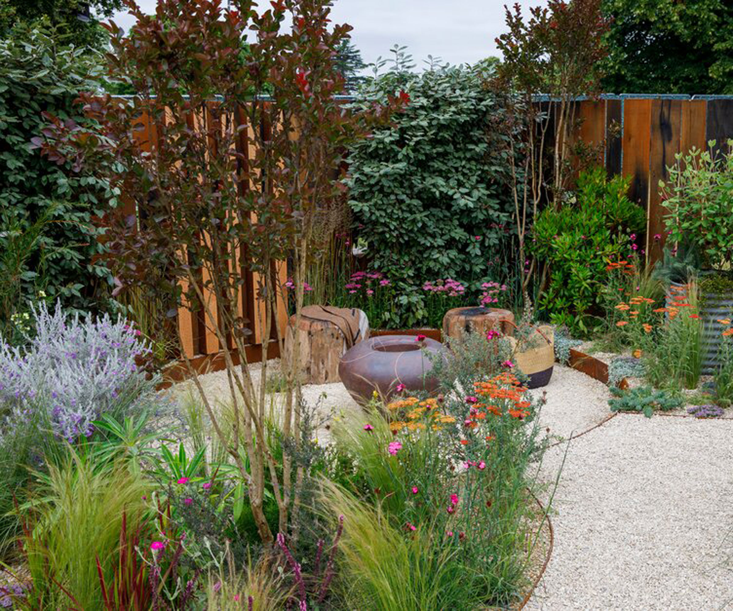 conception d'un petit jardin avec un parterre de fleurs profond comprenant des plantes, des fleurs, des graminées ornementales et un arbre à plusieurs tiges, un foyer et des sièges, une clôture et un aménagement paysager en gravier
