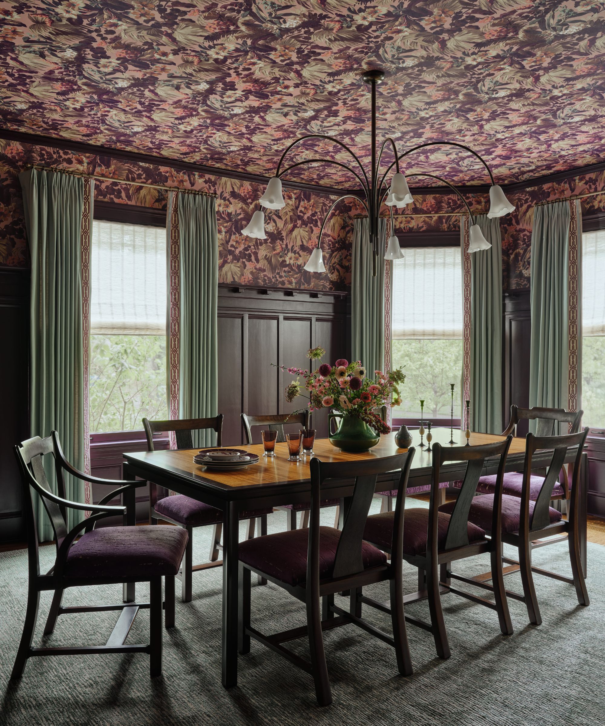 Salle à manger de mauvaise humeur avec des boiseries sombres, des chaises rembourrées violettes et un plafond assorti avec du papier peint floral foncé.