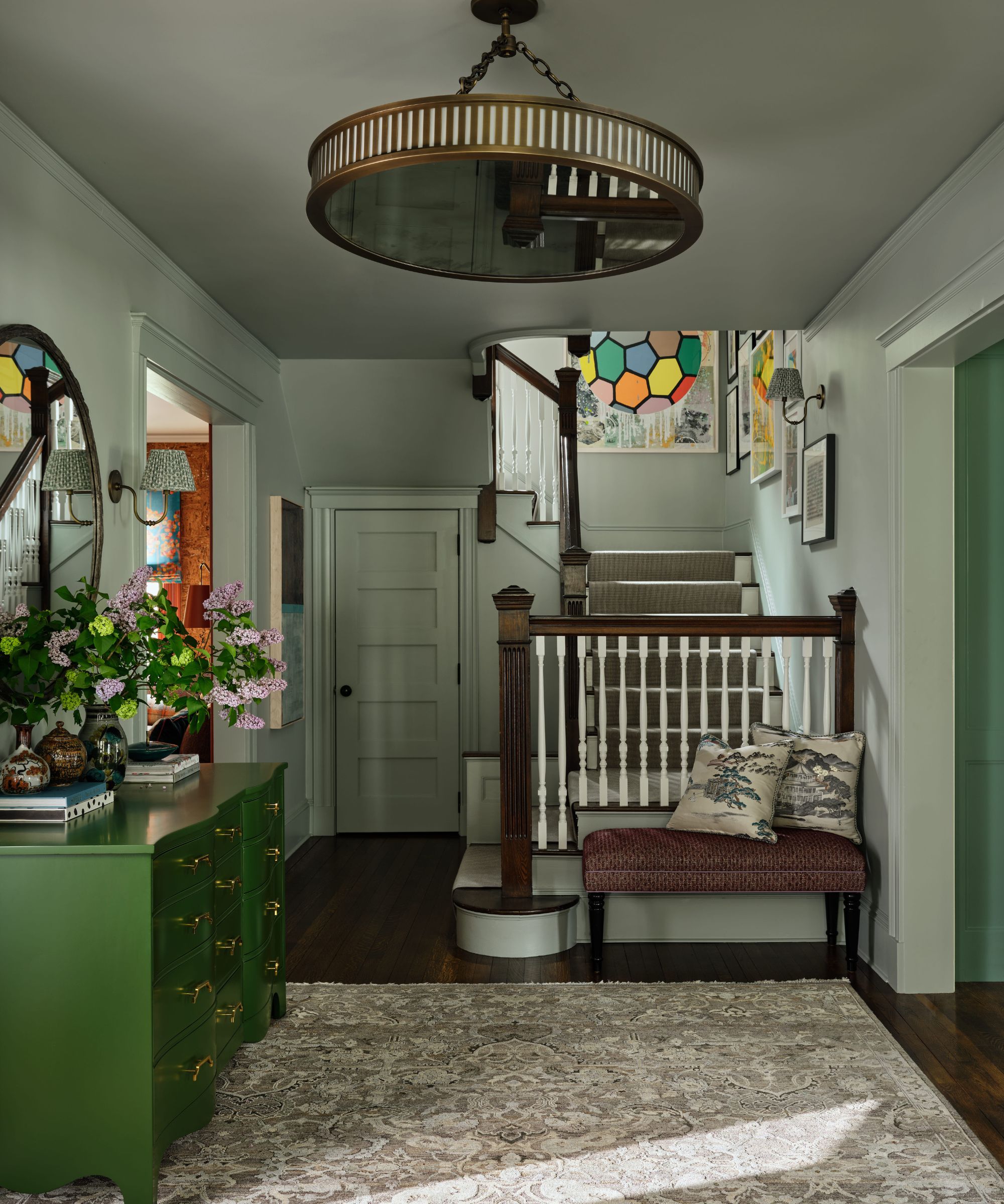 Entrée gris clair avec une commode verte, un escalier en bois avec une rampe rayée et un grand plafonnier rond en laiton.