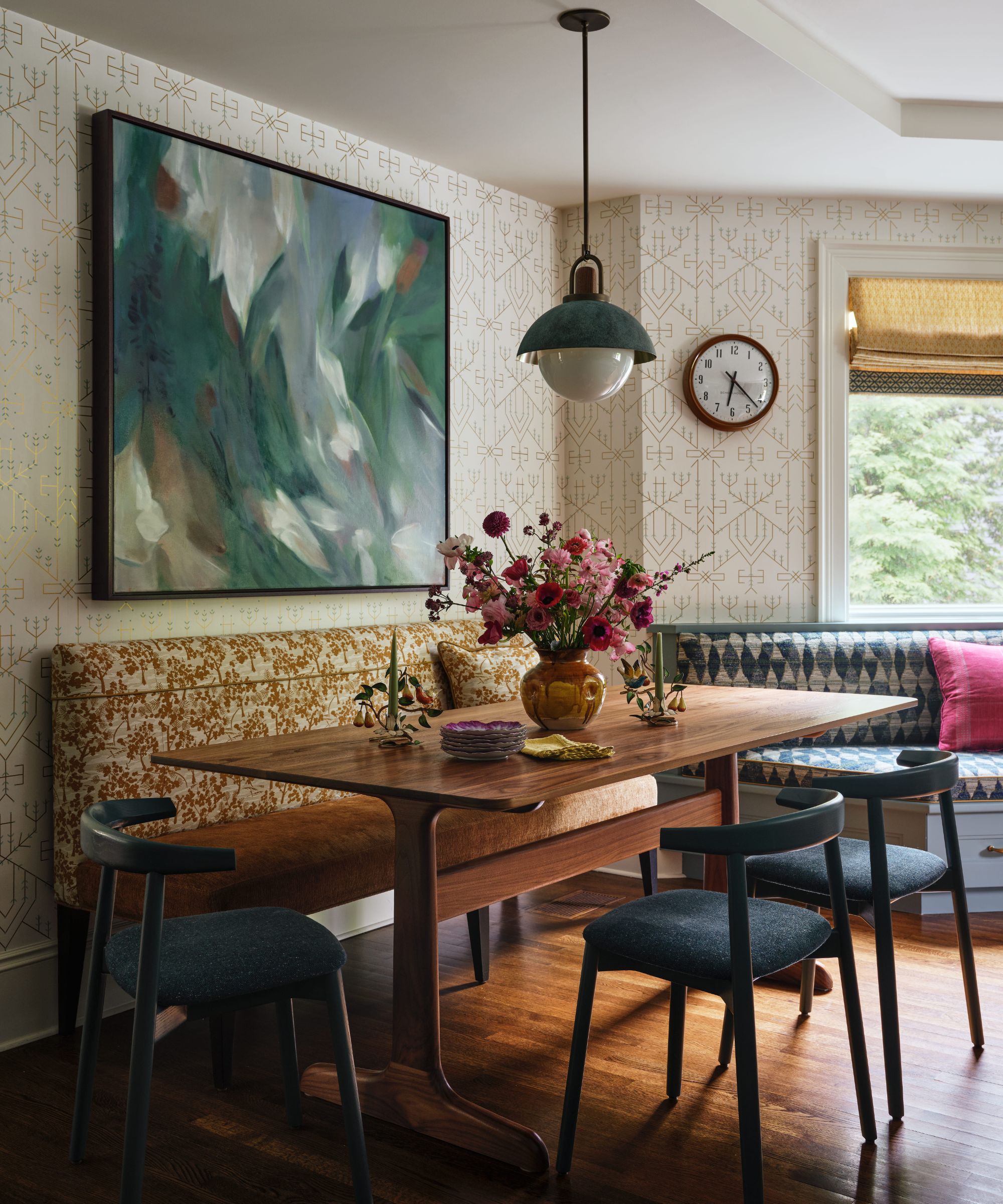 Coin repas comprenant une table en bois, des chaises sombres et un banc à motifs orange sous une grande peinture verte abstraite.