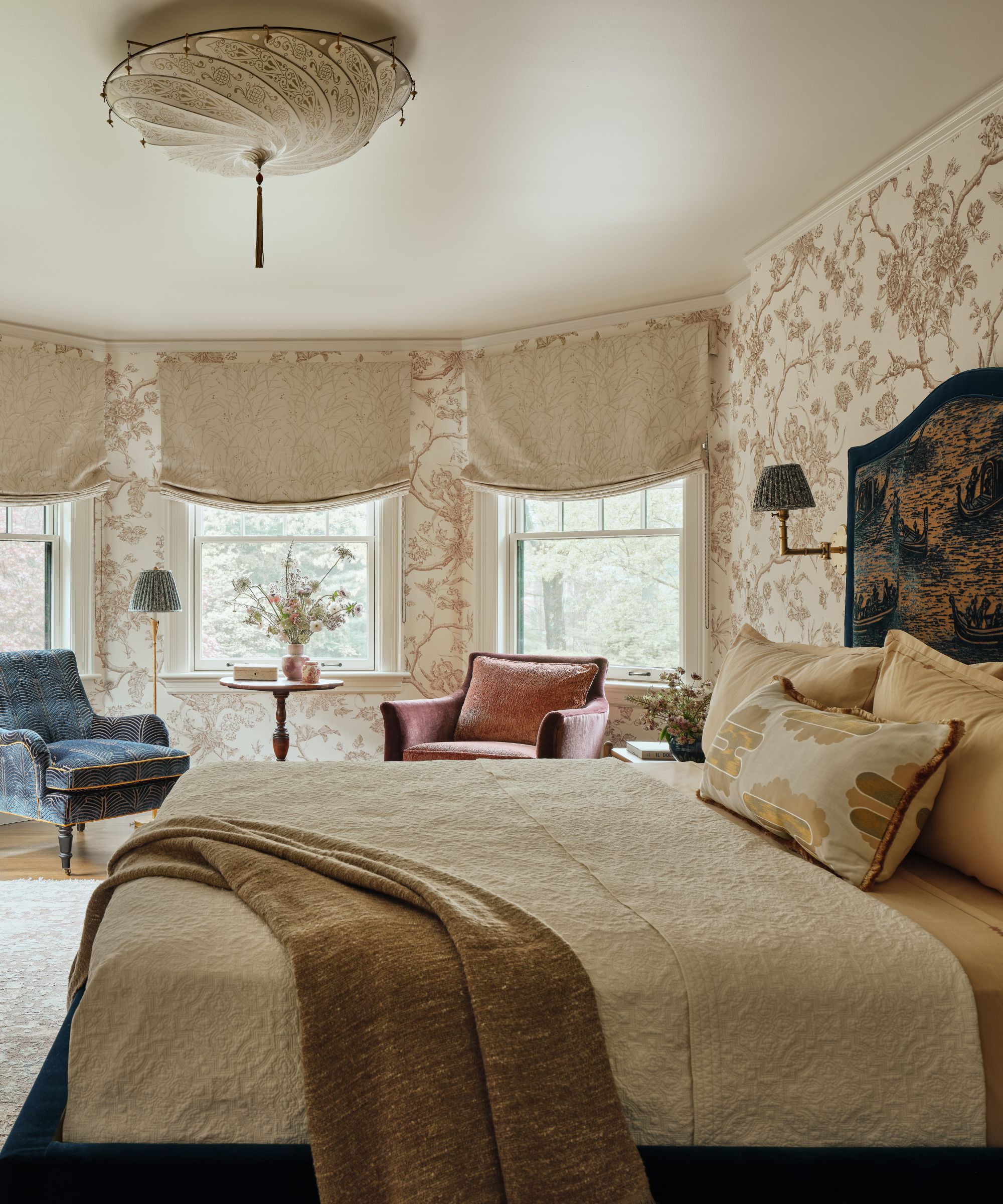 Chambre lumineuse avec un lit habillé de couleurs crème et marron, un papier peint floral neutre et deux fauteuils à motifs dans une baie vitrée.