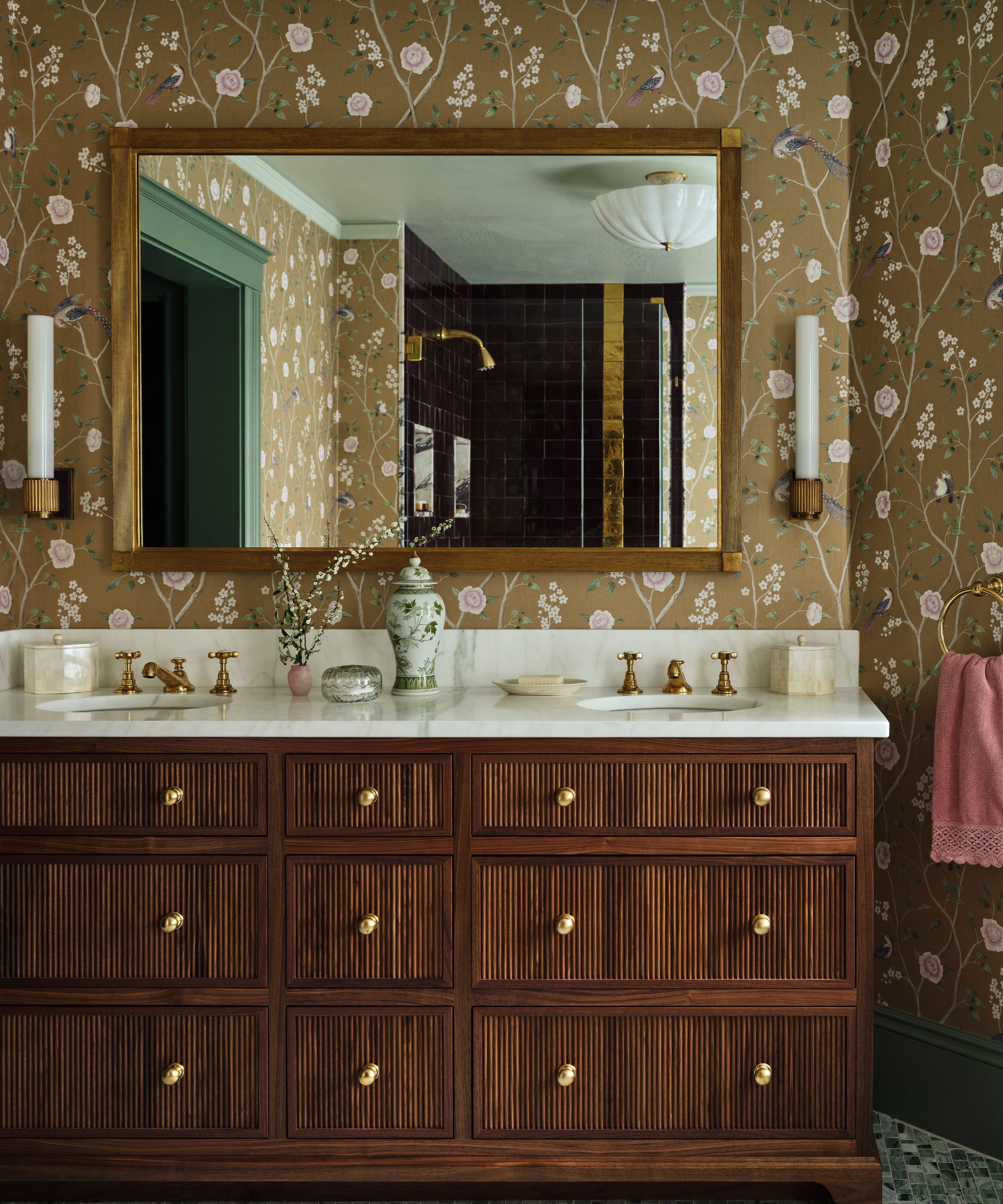 Meuble de salle de bain à double vasque avec armoires cannelées en bois foncé, dessus en marbre blanc et grand miroir au cadre doré sur fond de papier peint à fleurs marron