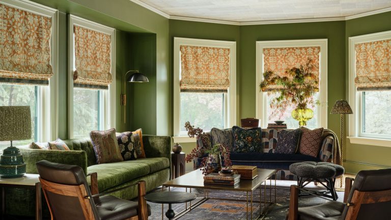 Cette maison de Boston des années 1800 était autrefois complètement grise – désormais, chaque pièce est remplie de couleurs vives et de couches de motifs A cozy living room with moss green walls, five windows with orange patterned blinds, white trim and ceiling, two velvet sofas and two matching leather and wood armchairs