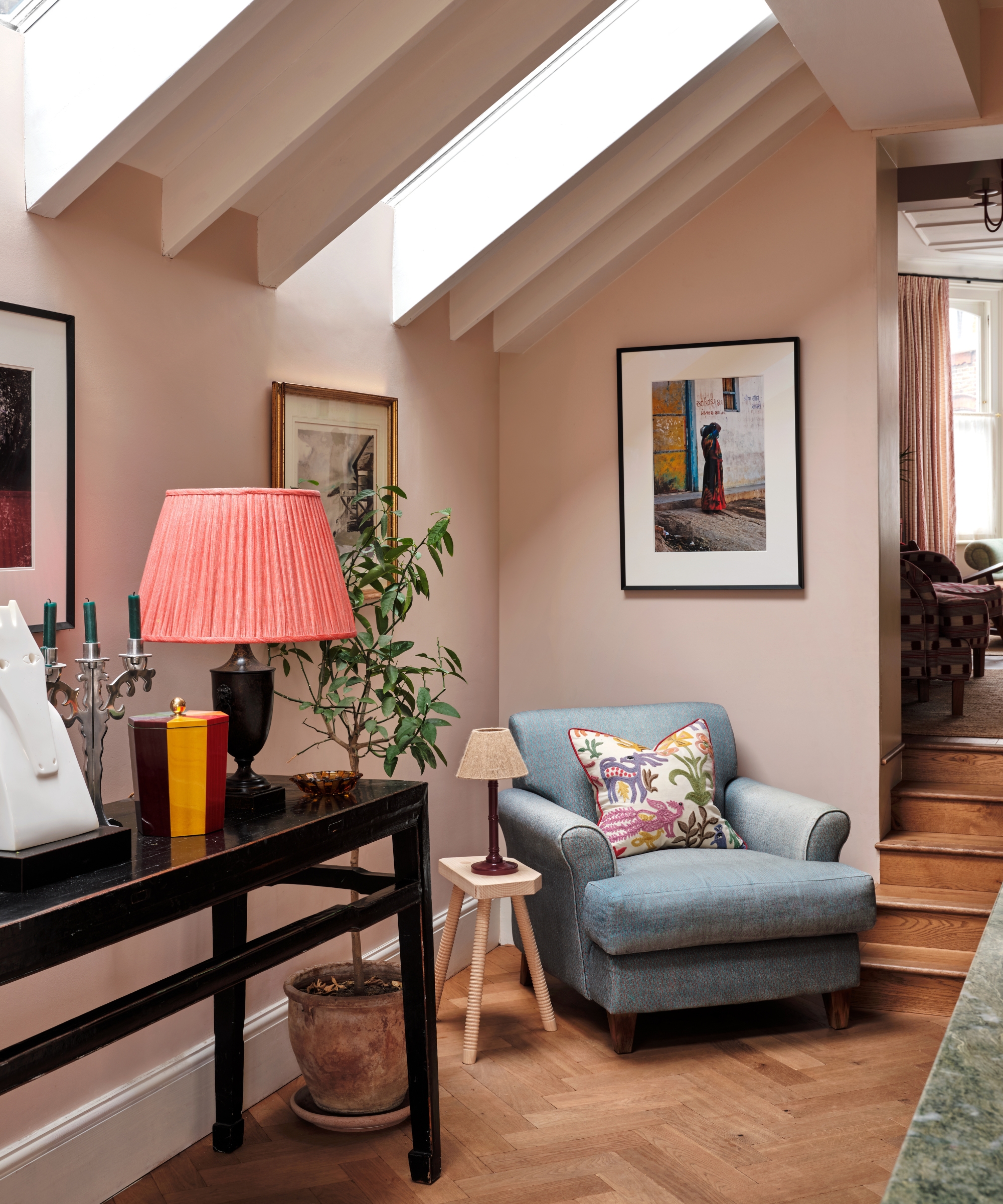 Le coin d'une cuisine aux murs roses et au parquet en chevrons, avec un fauteuil bleu, une table crédence et des œuvres d'art encadrées sur les murs