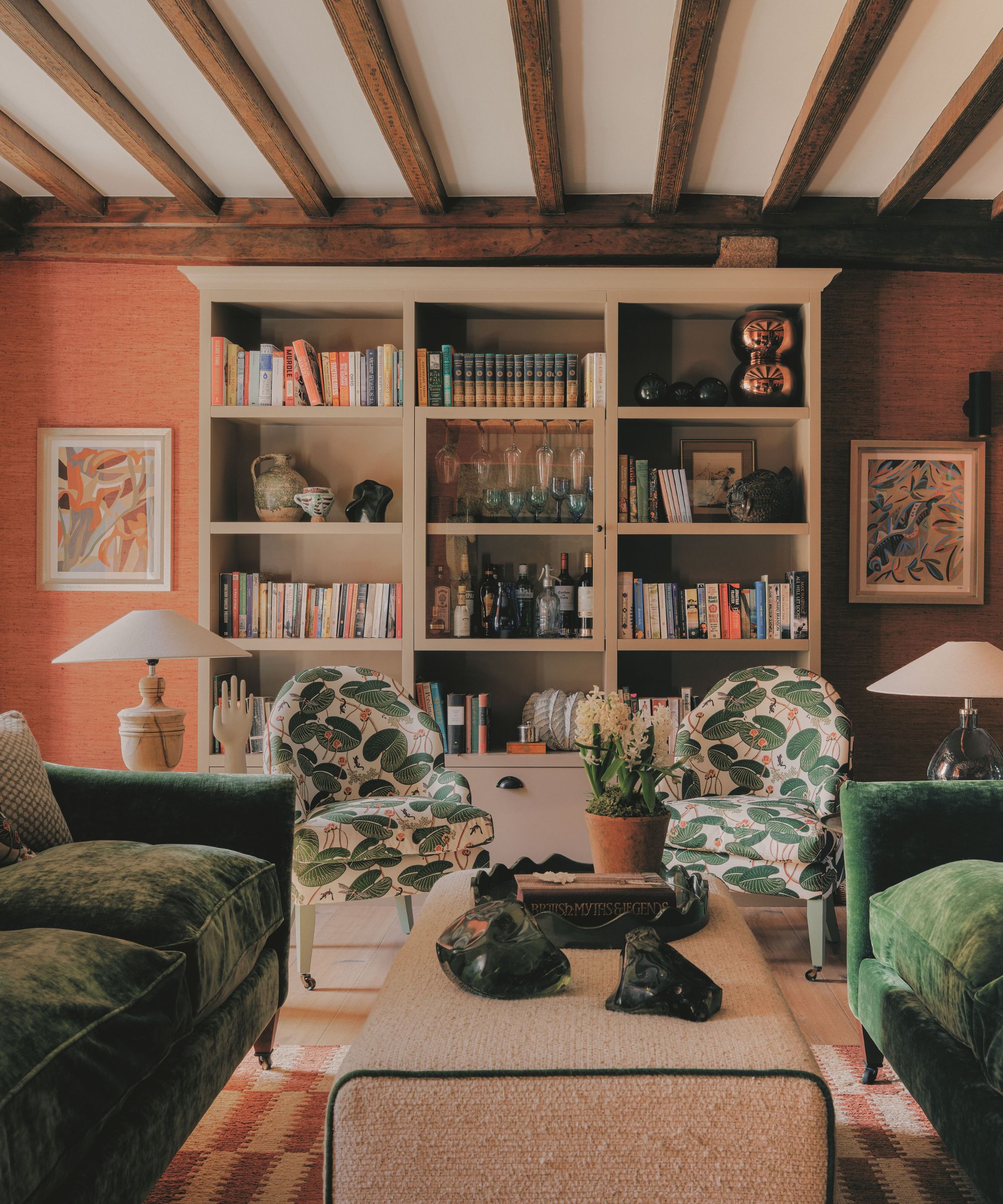 Vue du salon montrant des canapés en velours vert, des fauteuils à motifs, une table basse ottomane centrale, des poutres apparentes au plafond et des étagères remplies.