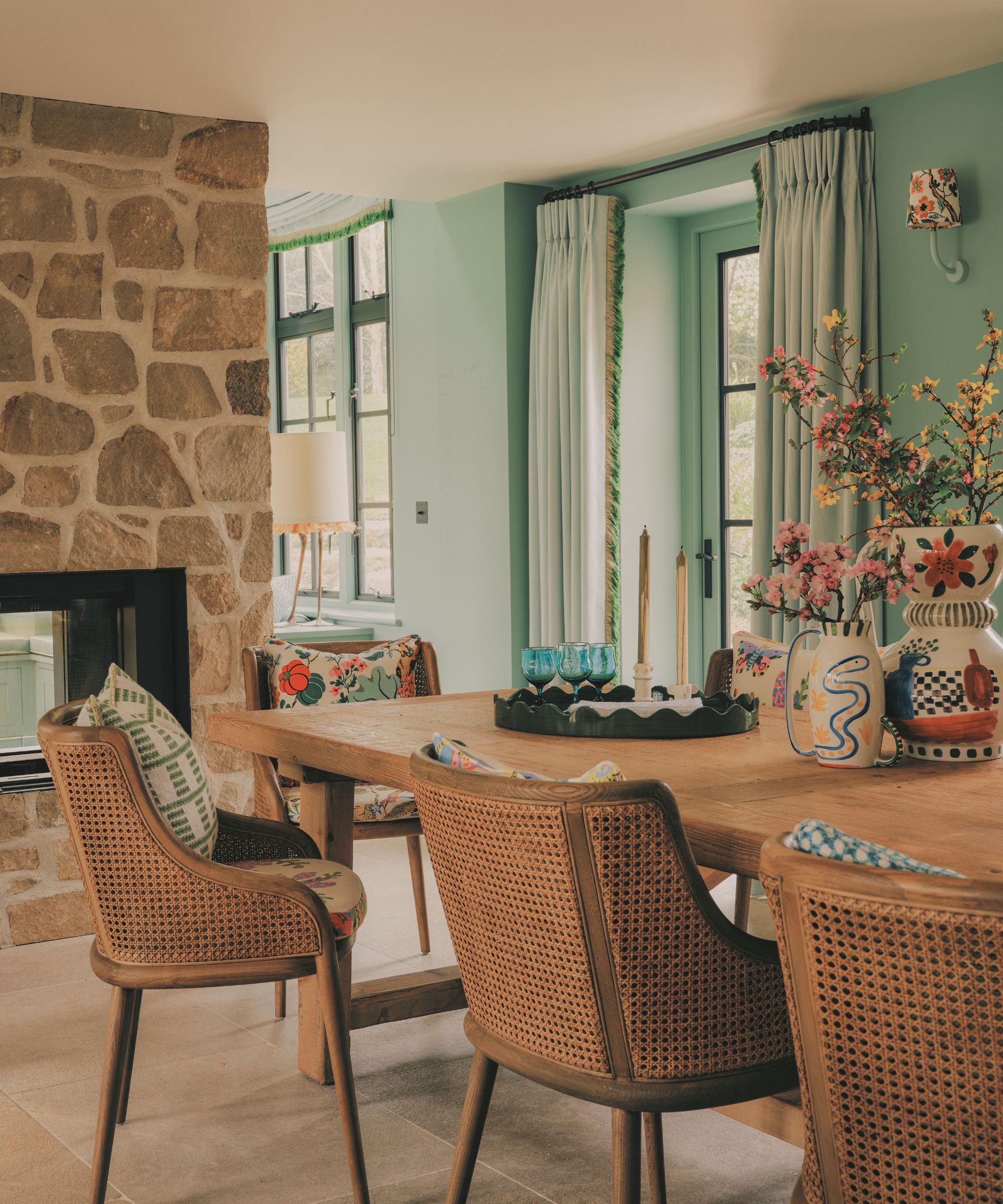 Salle à manger rustique avec chaises en rotin tressé autour d'une table en bois, murs vert menthe, cheminée en pierre et céramiques florales décoratives.