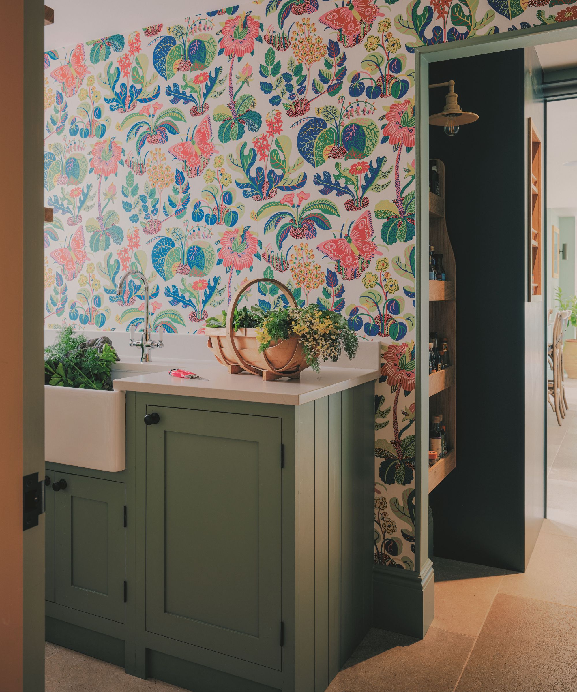 Espace utilitaire avec papier peint coloré à fleurs et papillons, armoires vert sauge, un évier blanc Belfast et un panier en bois rempli d'herbes fraîches.