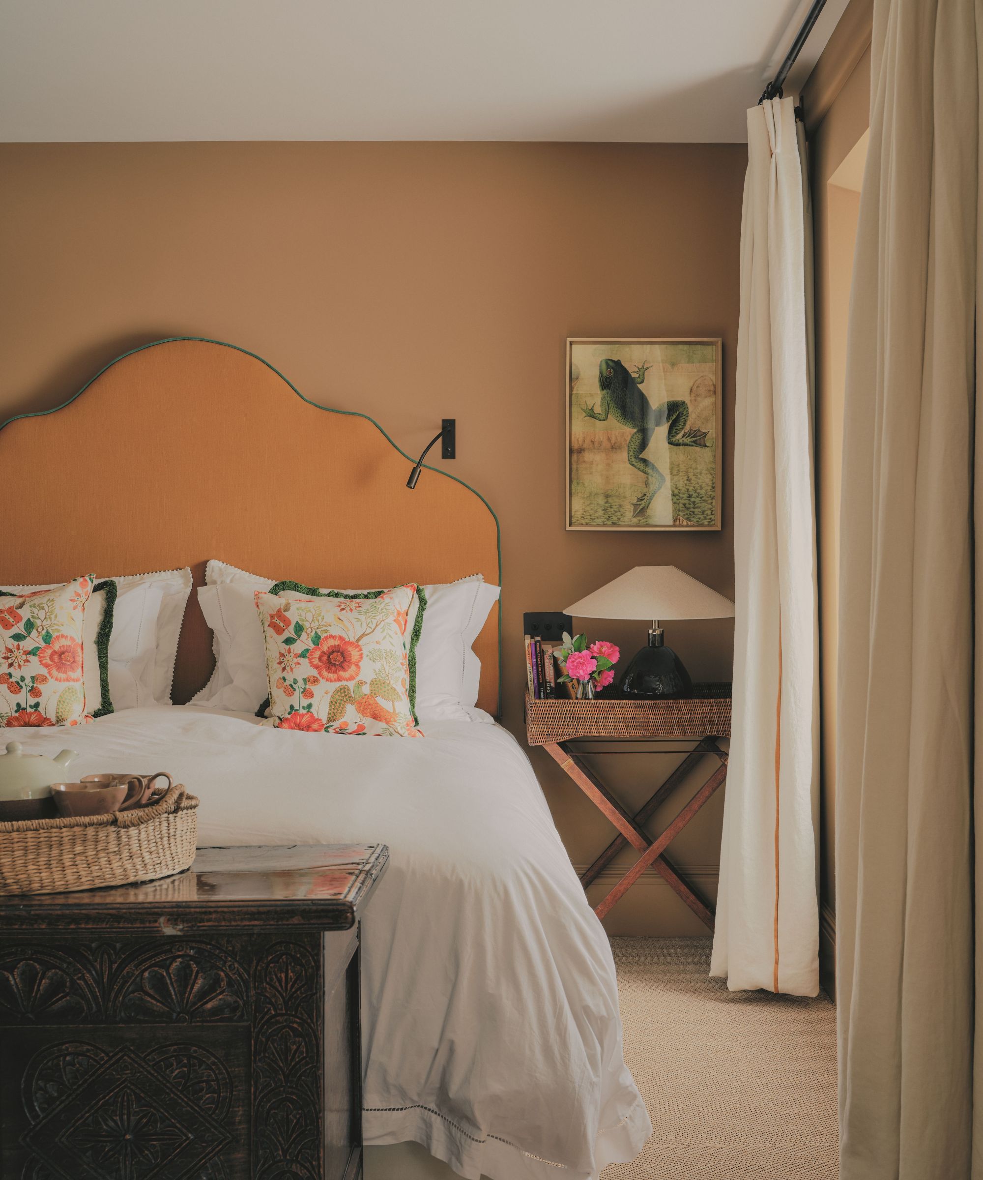 Chambre aux tons chauds comprenant une tête de lit incurvée orange, une literie blanche avec des oreillers aux accents floraux, une table de chevet avec une lampe et une œuvre d'art en forme de grenouille encadrée.