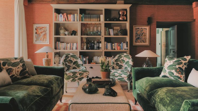 Cozy living room with terracotta walls, dark green velvet sofas, botanical patterned armchairs, and built-in bookshelves filled with books and glassware.