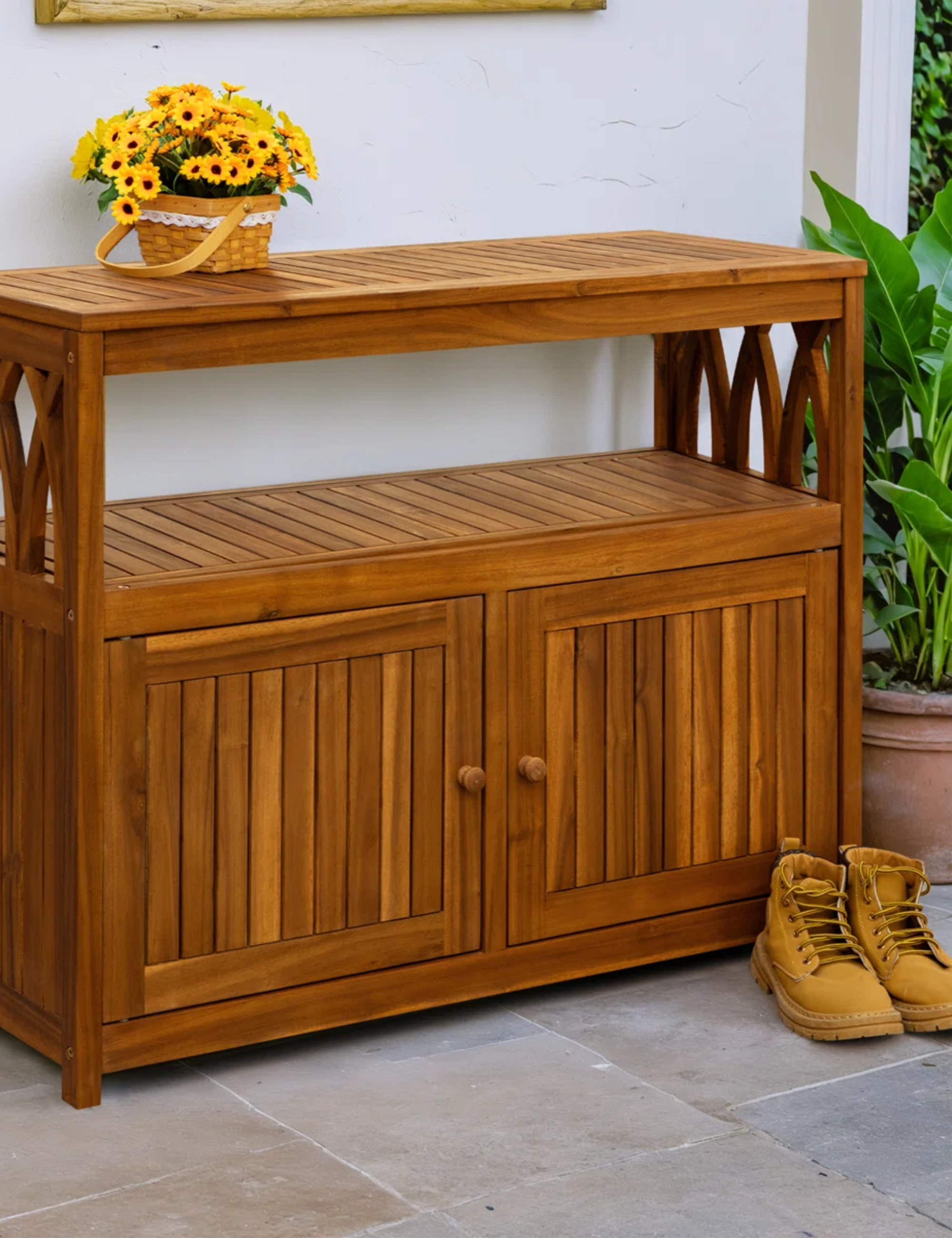 Buffet d'extérieur en bois décoré d'une paire de bottes de travail et d'un panier de tournesols au sommet d'un patio au sol en pierre.