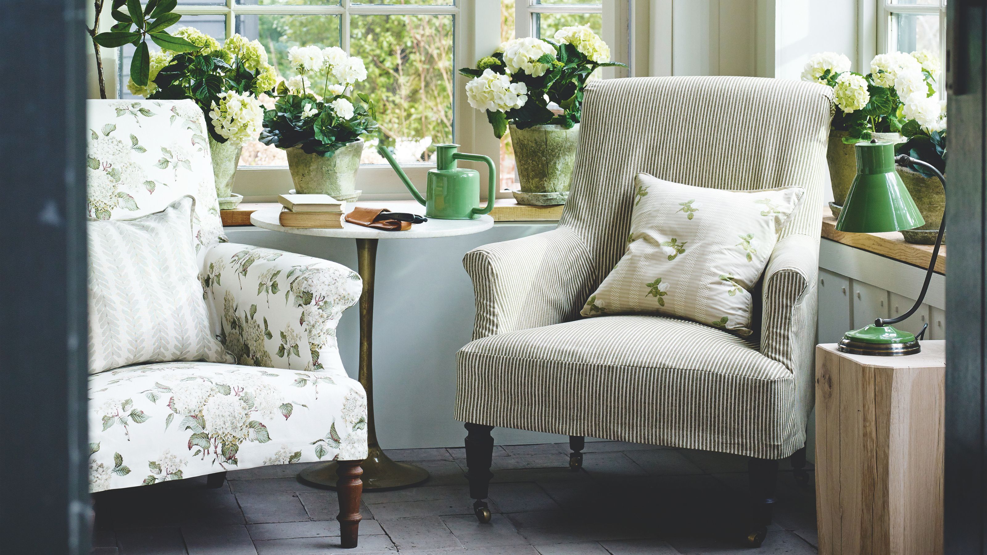 Coin salon dans une véranda ensoleillée avec carrelage au sol, un fauteuil fleuri à côté d'un fauteuil rayé, des fleurs blanches, un arrosoir vert sur une table circulaire et une lampe verte, entourée de fenêtres.