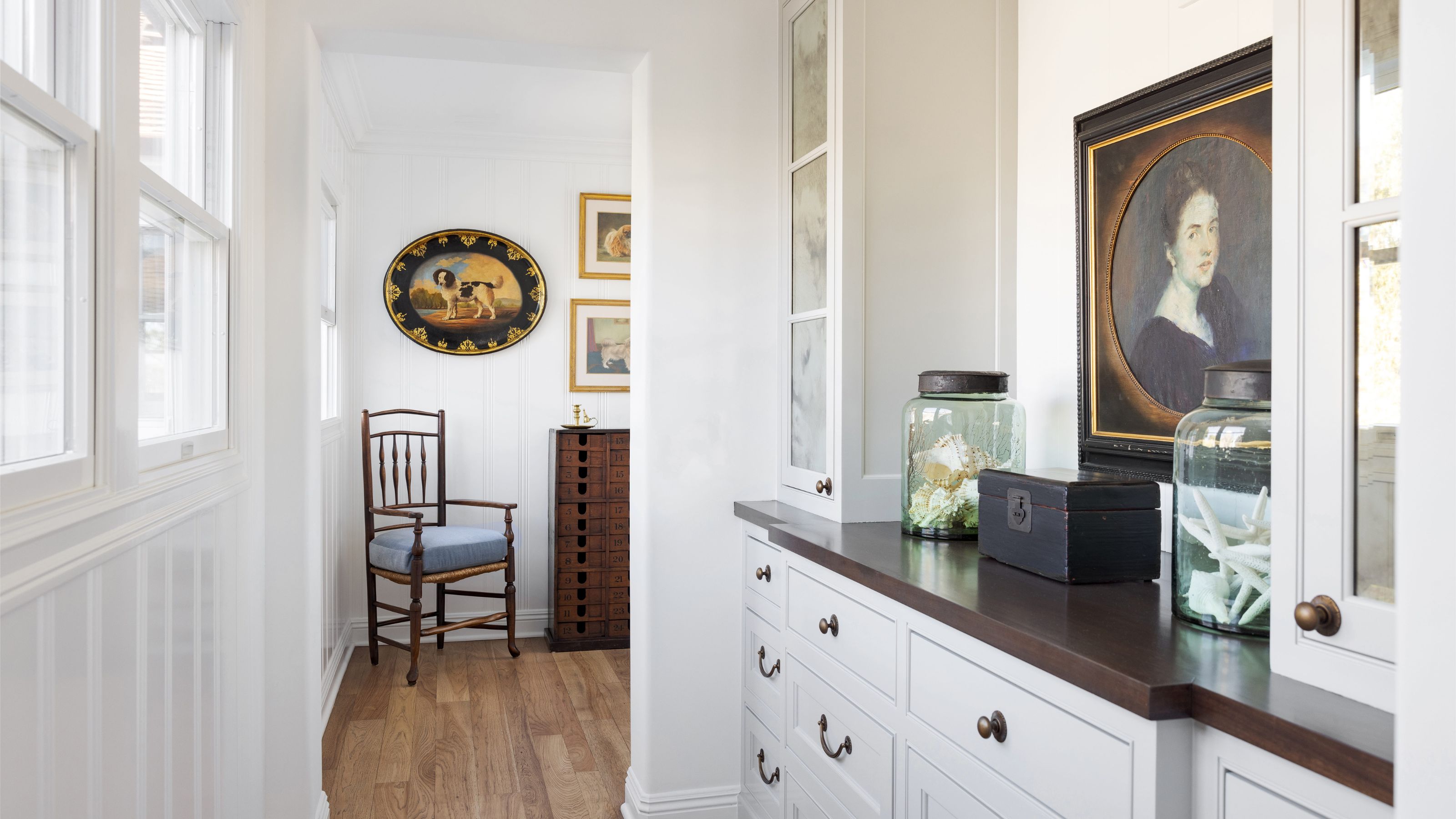 Cuisine américaine avec armoires blanches, comptoirs noirs, armoires vitrées, portrait antique, bocaux en verre remplis de coquillages et chaise en bois vintage surmontée d'œuvres d'art vintage.