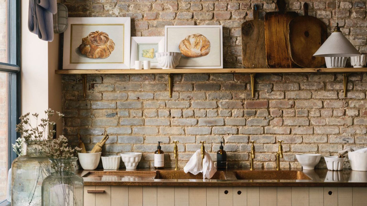 Pourquoi les designers disent que chaque cuisine a besoin d’œuvres d’art – et comment trouver le juste équilibre urban kitchen with exposed brick wall, copper countertop, brass hardware, plywood cabinets, single open shelf with objects, artwork, white tableware, deVOL