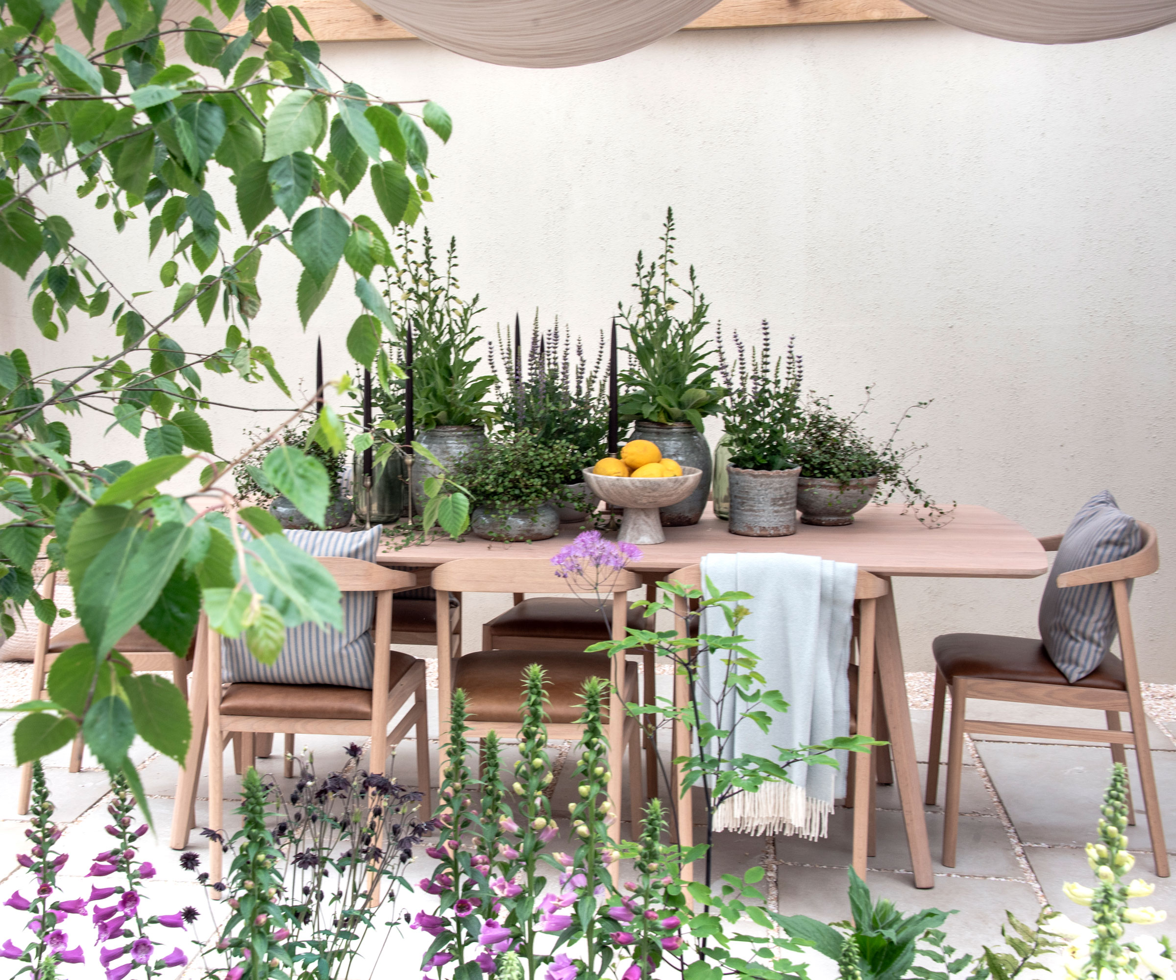 terrasse de jardin avec tables et chaises pour manger en plein air, avec des accessoires tels que des coussins et des plaids, des plantes en pot et des bougeoirs, et des digitales plantées dans un parterre de fleurs